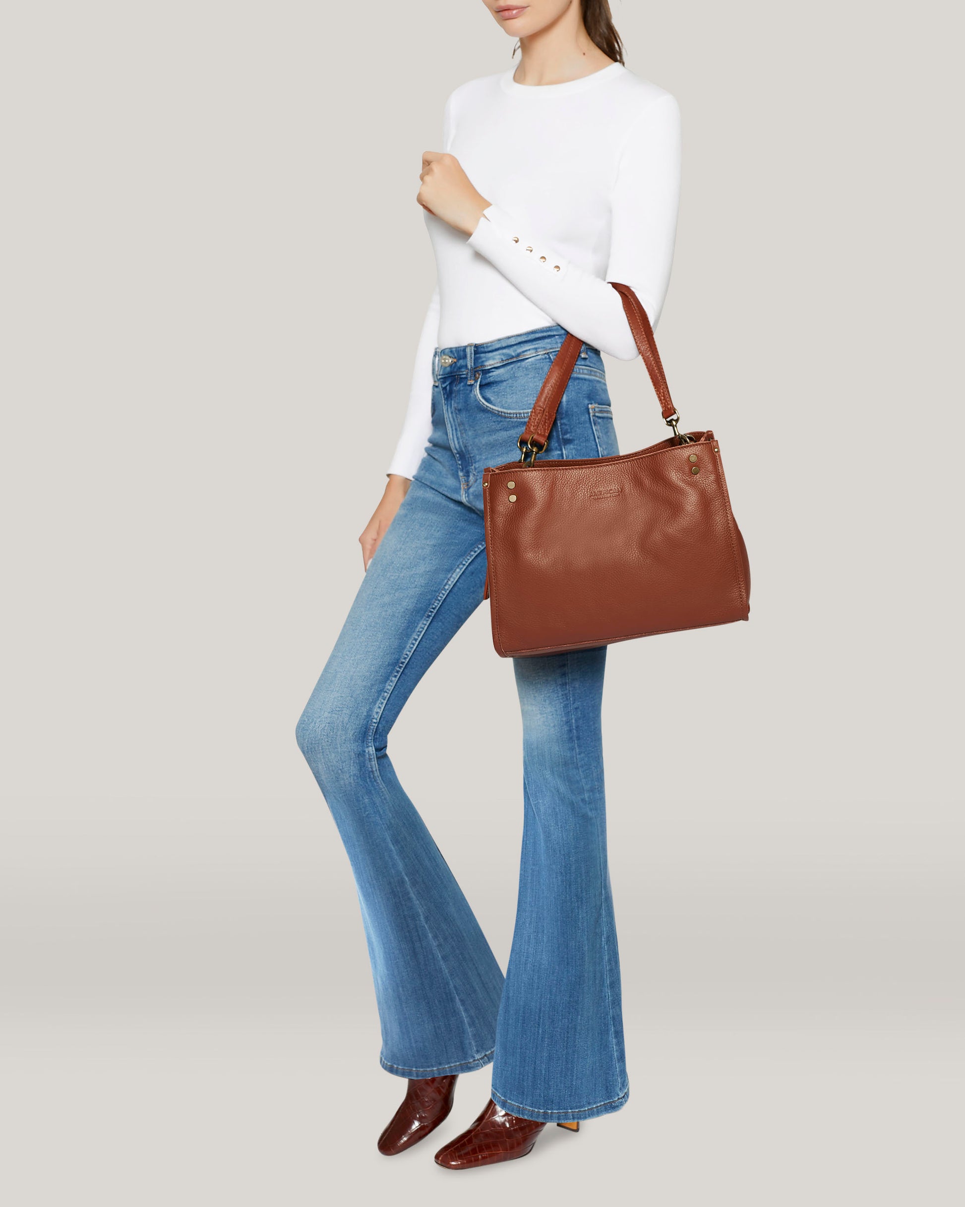 A person wearing a white long-sleeve shirt and blue flared jeans holds the chic Lenox Triple Entry Satchel from American Leather Co., crafted from genuine leather. Complemented by brown ankle boots against a light gray background, the ensemble is both stylish and functional with its thoughtfully designed interior compartments.