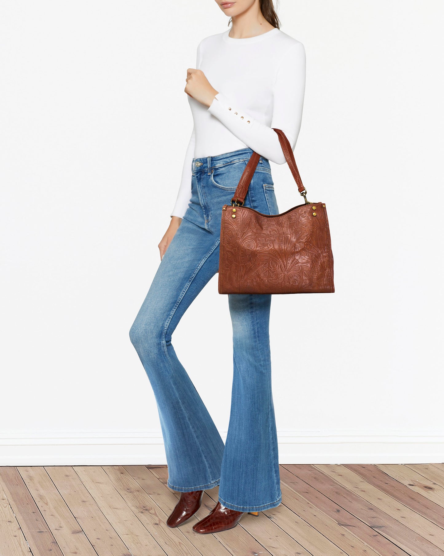 A person dressed in a white long-sleeve shirt and blue flared jeans stands on a wooden floor, holding an everyday essential: the Lenox Triple Entry Satchel by American Leather Co., a brown embossed leather handbag with dual handles. The person completes their look with brown boots against a plain white backdrop.