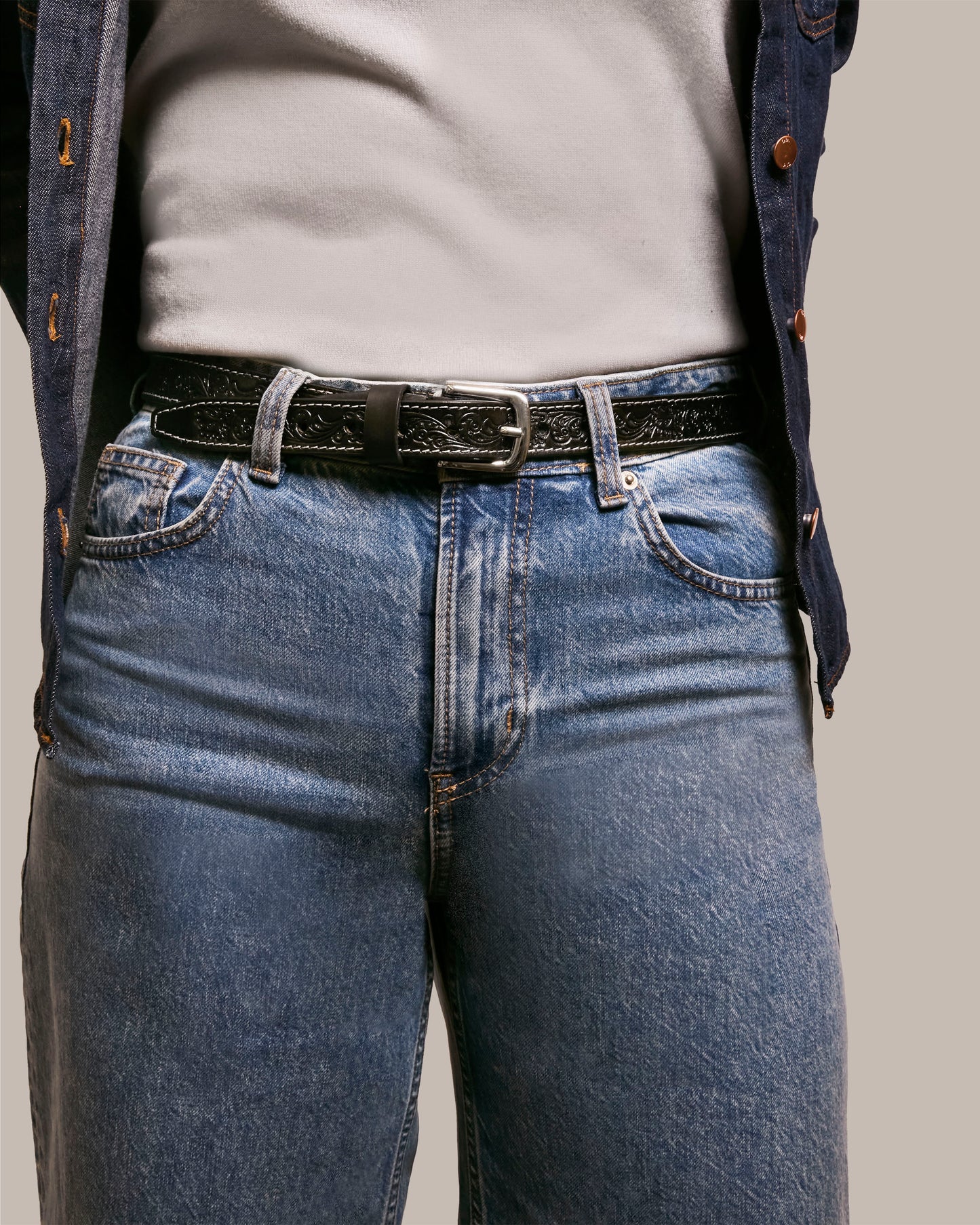 A person in faded blue jeans and a Lockport Belt by American Leather Co., with a silver buckle, wears a white shirt under a dark jacket. Their arms hang at their sides, drawing attention to the outfit's midsection details.