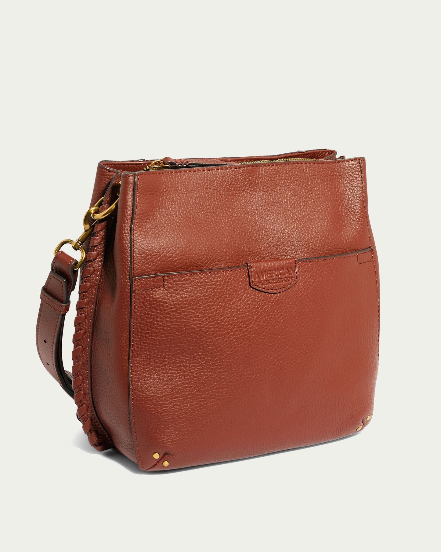 The Luca Triple Entry Crossbody by American Leather Co. is a brown leather bag featuring a shoulder strap, brass hardware details, a front pocket, decorative stitching along the sides, and a small embossed logo on the front. The background is a plain light color.