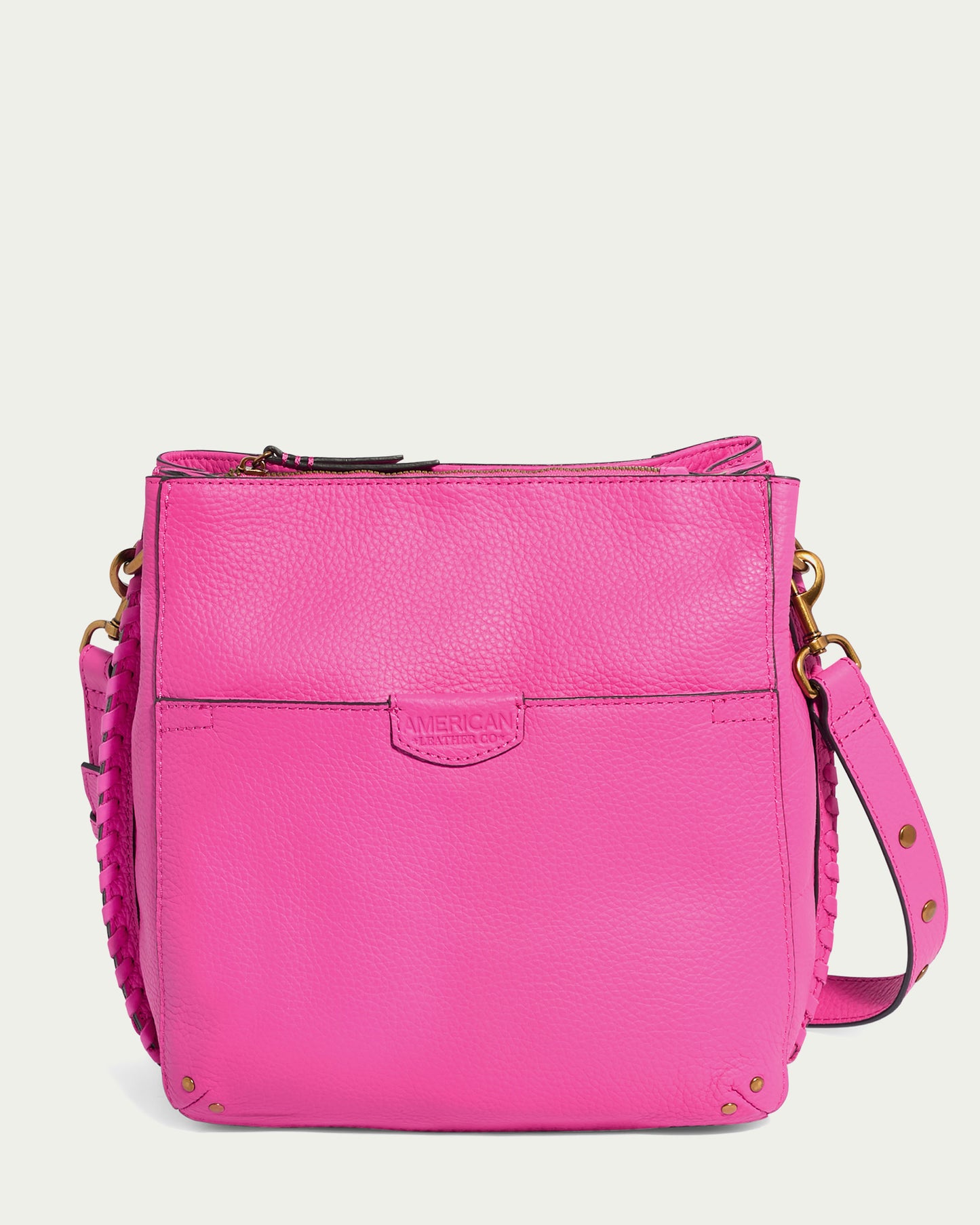 Introducing the Luca Triple Entry Crossbody by American Leather Co.: a bright pink leather bag made from glove-tanned pebble leather, featuring gold hardware. It boasts a front pocket, zippered main compartment, braided accents on the adjustable strap adorned with gold rivets, and an embossed logo on the front.