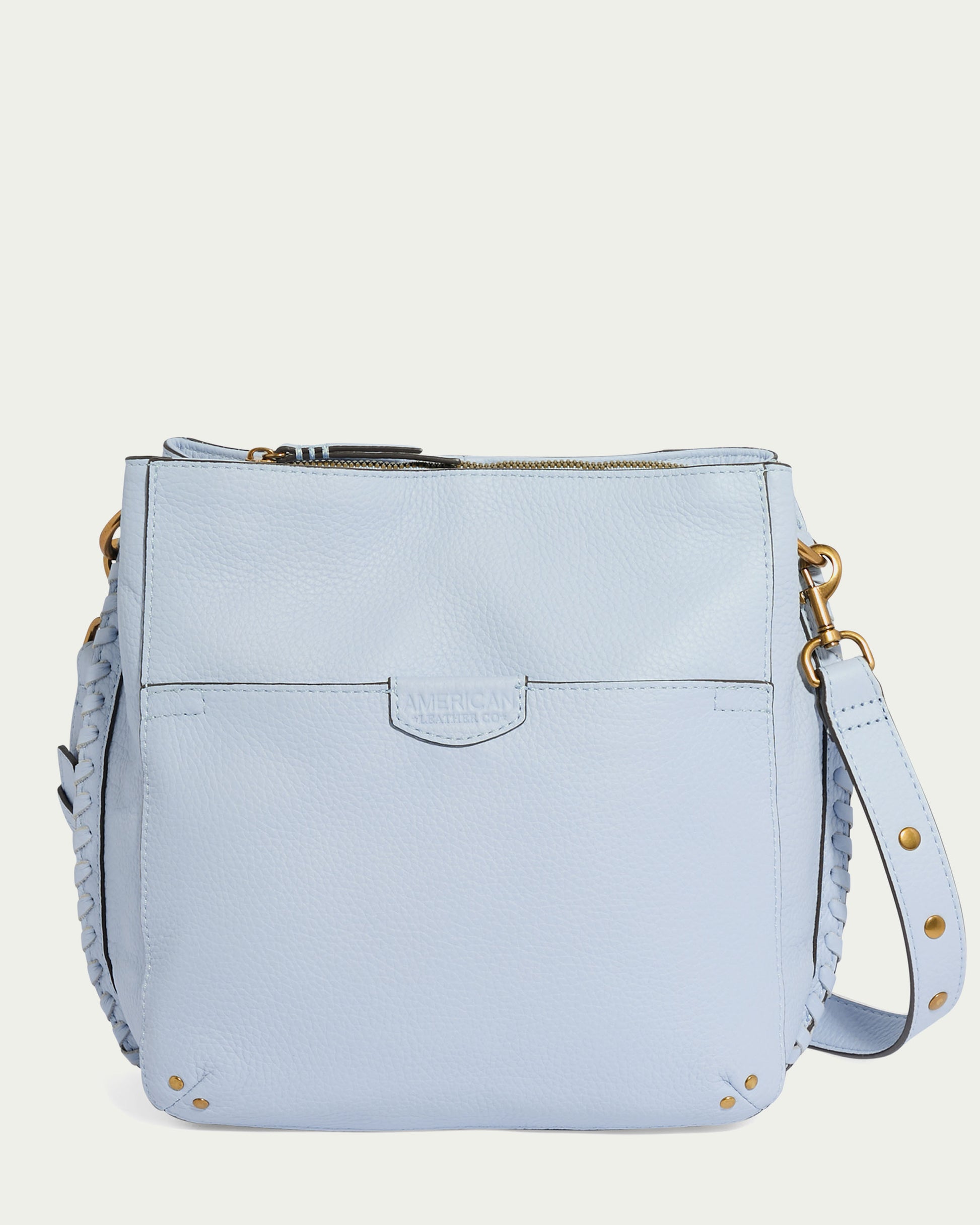 A light blue leather shoulder bag featuring a zippered top closure, a front pocket, brass hardware, and a braided adjustable/removable crossbody strap with brass rivets. Made from glove-tanned pebble leather, the Luca Triple Entry Crossbody by American Leather Co. has the brand name subtly embossed on the front pocket.