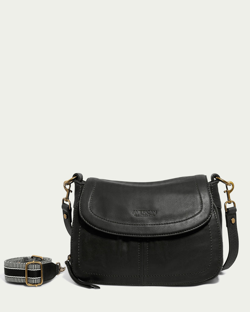 The Marino Crossbody with 2 Straps by American Leather Co. is a black leather shoulder bag adorned with a flap closure and brass hardware, with the brand name embossed on the front. It boasts an adjustable, removable strap featuring a distinctive black and white geometric design, highlighting its functional design crafted from premium leather.