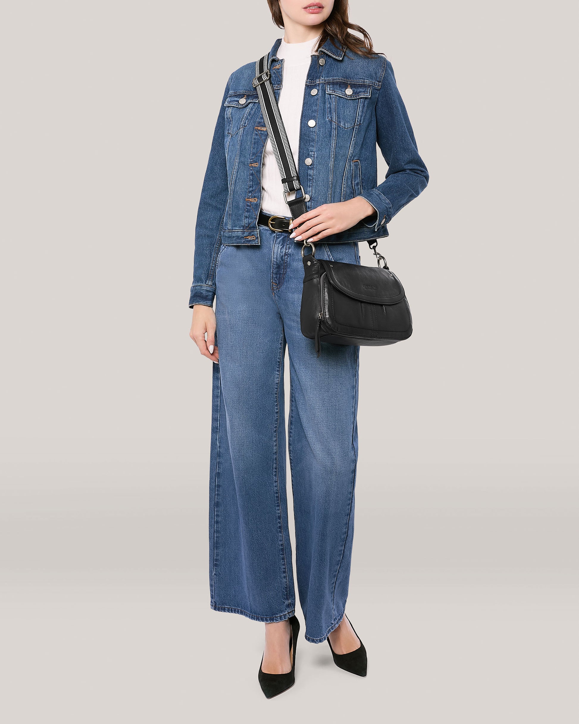 A person wearing a denim jacket over a white top, wide-leg denim jeans, and black heels stands against a plain background. They are holding an American Leather Co. Marino Crossbody with 2 Straps in butter-soft leather, draped across their body.