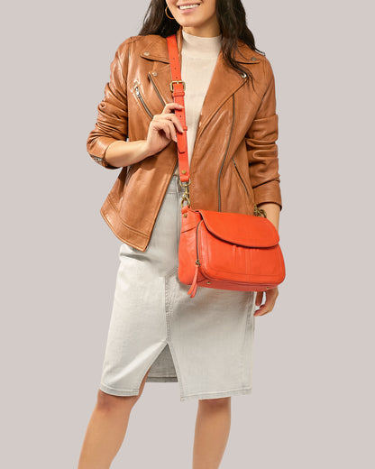 A person wearing a brown leather jacket, a light-colored skirt, and a beige top is holding an American Leather Co. Marino Crossbody with 2 Straps in bright orange. The background is plain and light gray. The outfit has a casual, stylish look, perfectly complemented by the bag's premium leather and functional design.