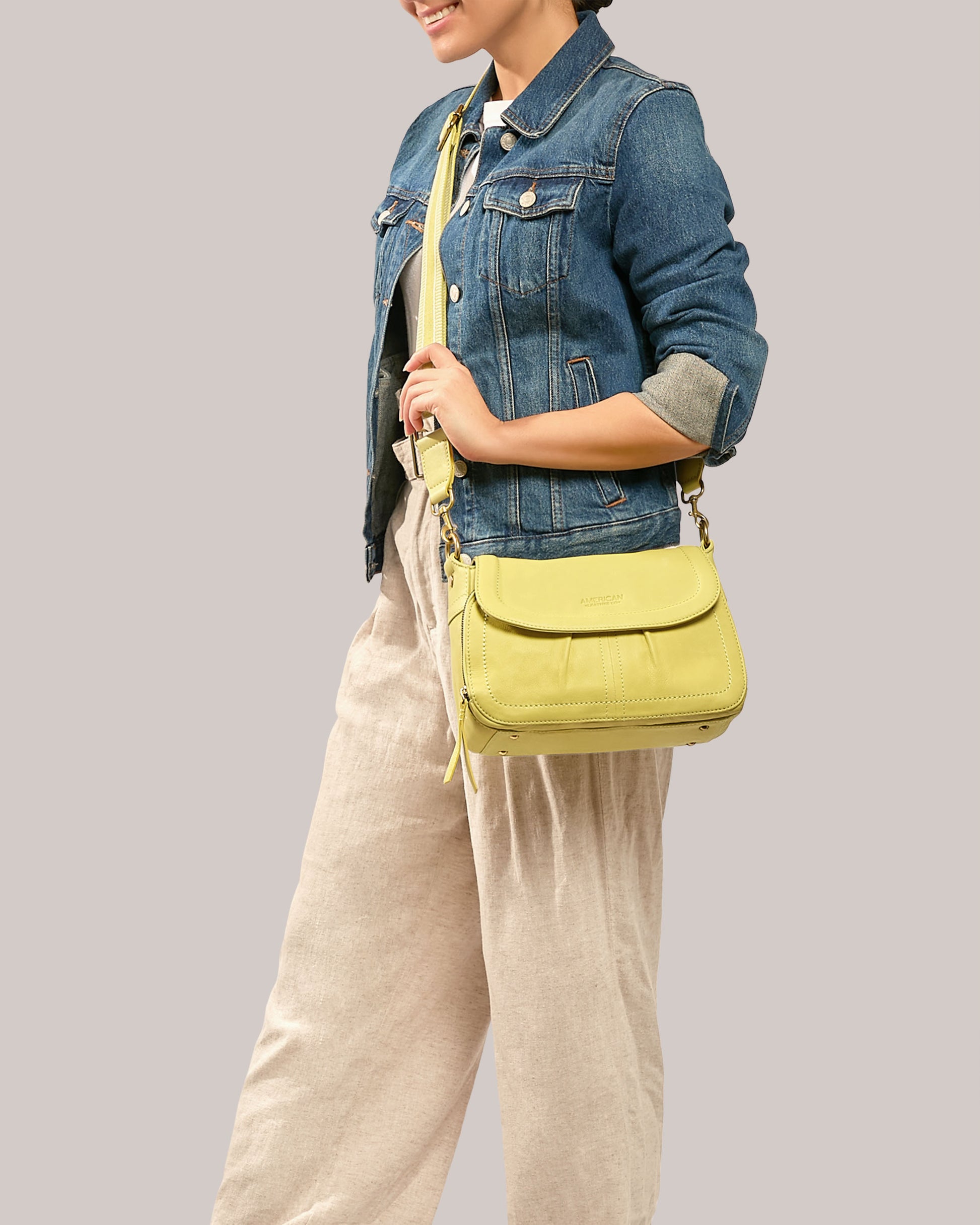 A person wearing a denim jacket and light beige pants holds an American Leather Co. Marino Crossbody with 2 Straps, crafted from premium leather. The background is plain gray.