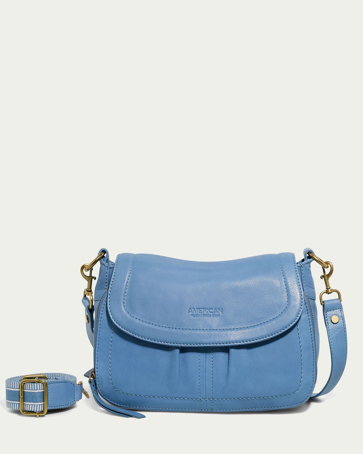 A light blue Marino Crossbody with 2 Straps, crafted from butter-soft leather with a flap closure and gold-tone hardware. It features a detachable strap with an adjustable gold buckle, along with a front pocket. The brand "American Leather Co." is elegantly embossed on the flap, accentuating its functional design.