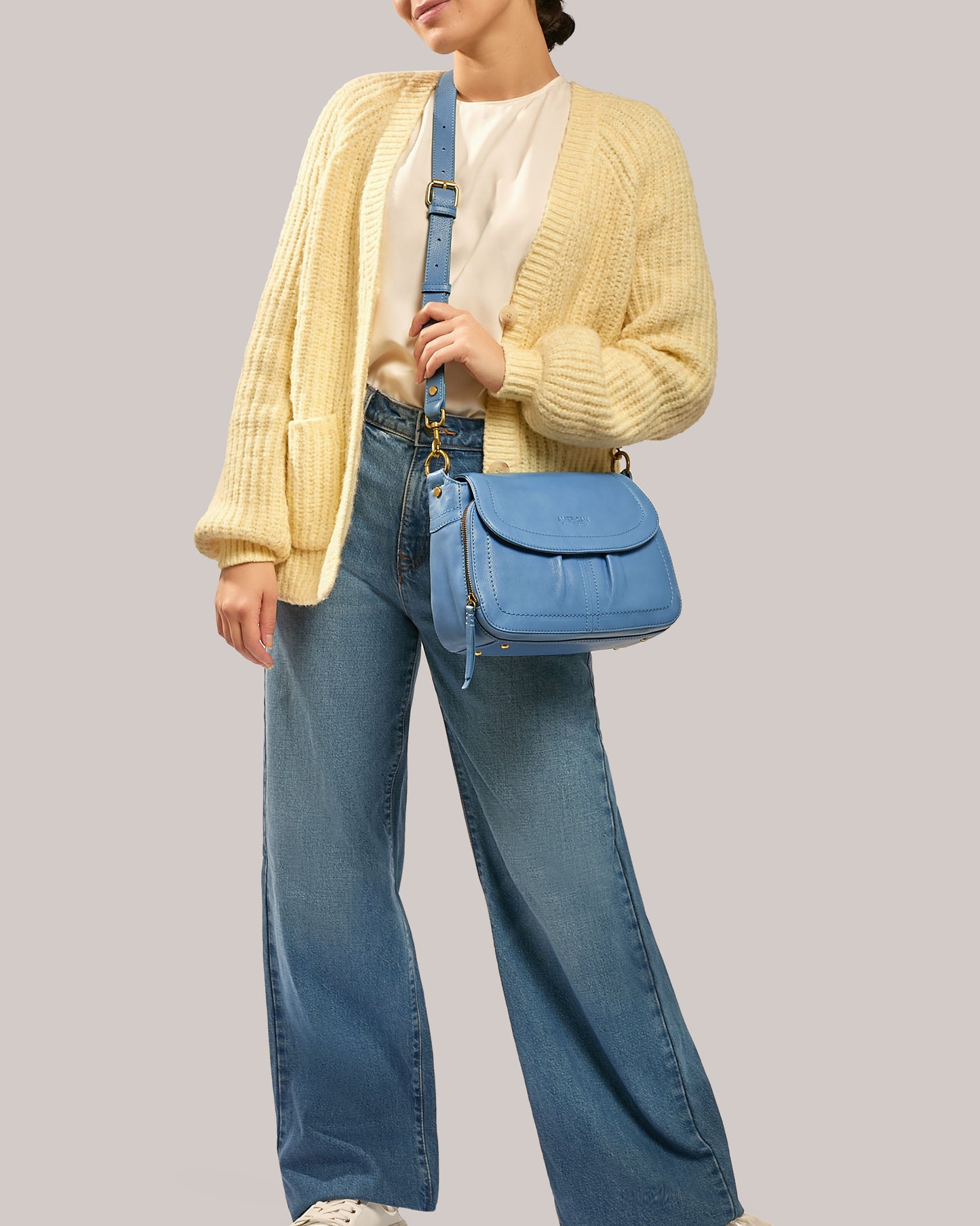 Dressed in a cream cardigan, white blouse, and blue wide-leg jeans, this individual carries the vibrant Marino Crossbody with 2 Straps by American Leather Co. in butter-soft leather. The ensemble is perfectly completed with white sneakers against a plain, neutral backdrop.