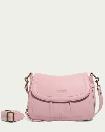 A pastel pink Marino Crossbody with 2 Straps, featuring a flap closure, is displayed against a light background. The butter-soft leather bag boasts brass hardware and comes with a removable, adjustable shoulder strap. The brand name "American Leather Co." is embossed on the front flap.