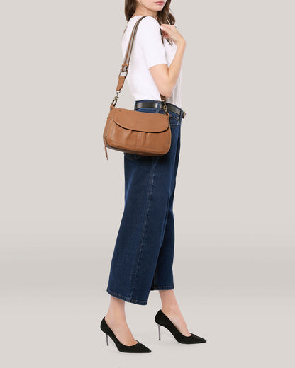 A woman in a white t-shirt, blue high-waisted jeans, and black high-heeled shoes stands against a plain background. She carries an American Leather Co. Marino Crossbody with 2 Straps in butter-soft leather featuring an adjustable, removable strap. Her head is turned to the side, and her left hand is near her chin.