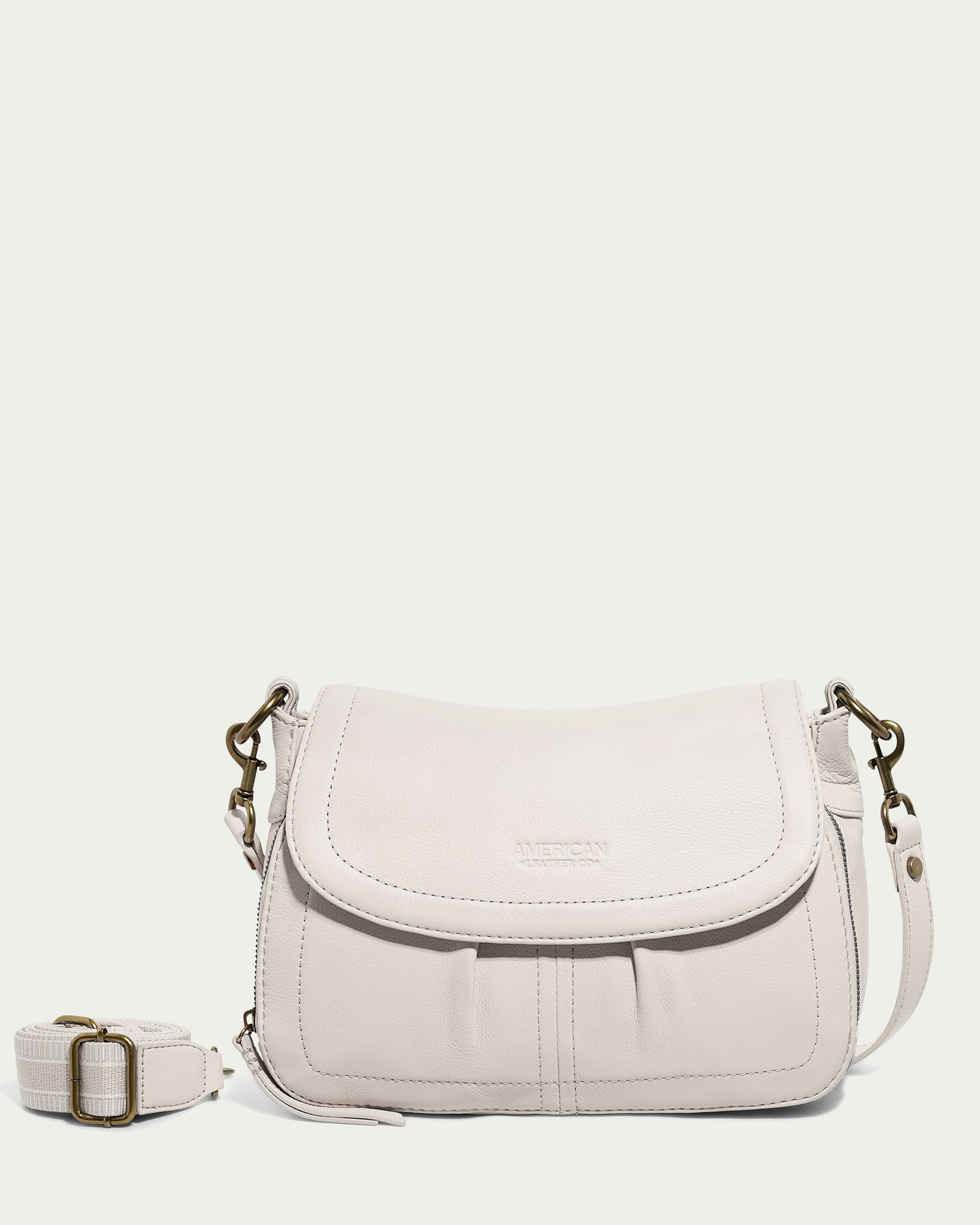 A light beige leather Marino Crossbody with 2 Straps from American Leather Co. features a flap closure and a minimalistic design. The flap includes subtle stitching and the brand name embossed at the center. An adjustable, detachable shoulder strap with a metallic clip positioned to the left of the bag enhances its functional design.