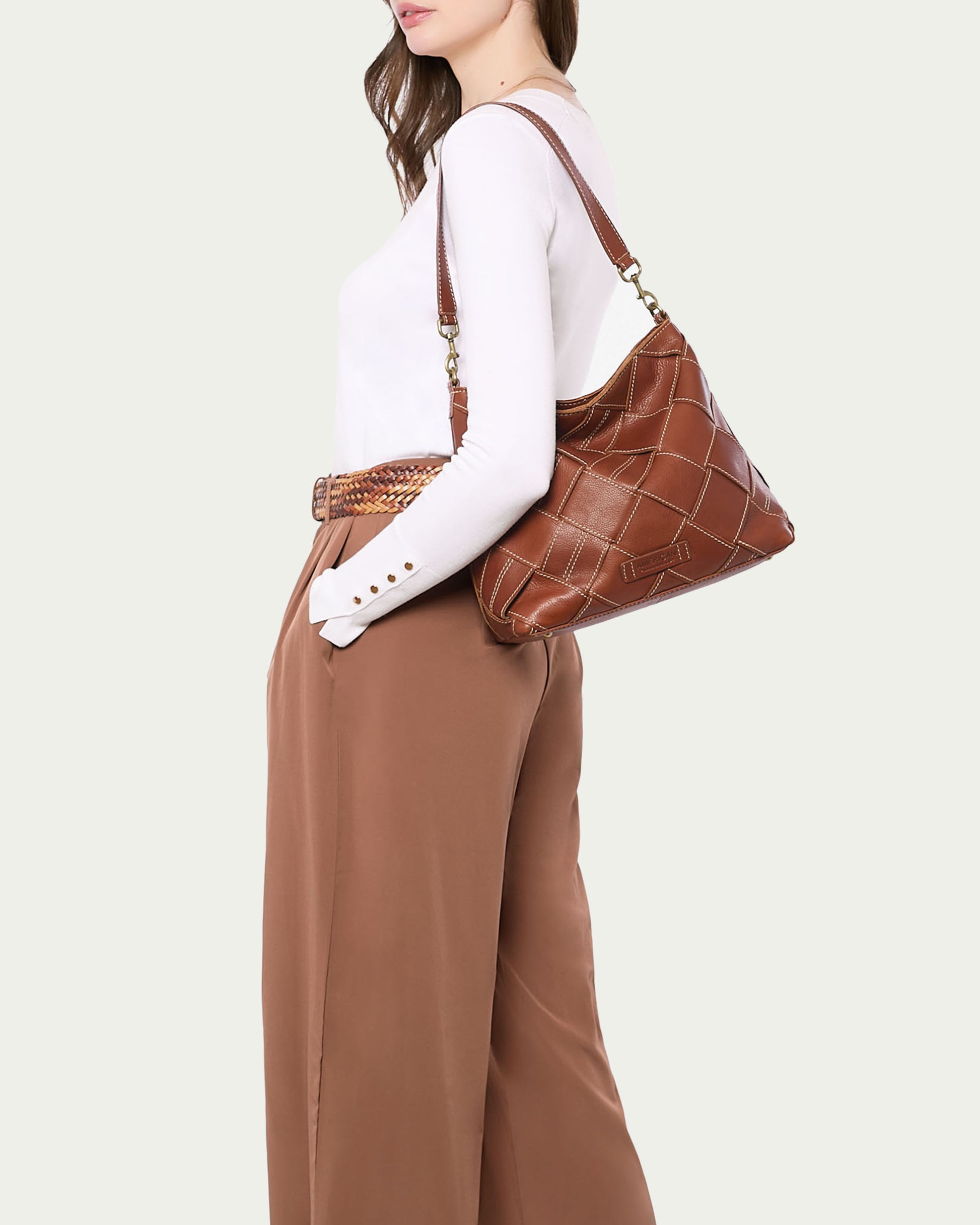 Person wearing a white long-sleeve top and brown high-waisted pants carries an American Leather Co. Mercer Woven Top Zip Hobo bag with a removable strap, elegantly accessorized with a patterned belt.