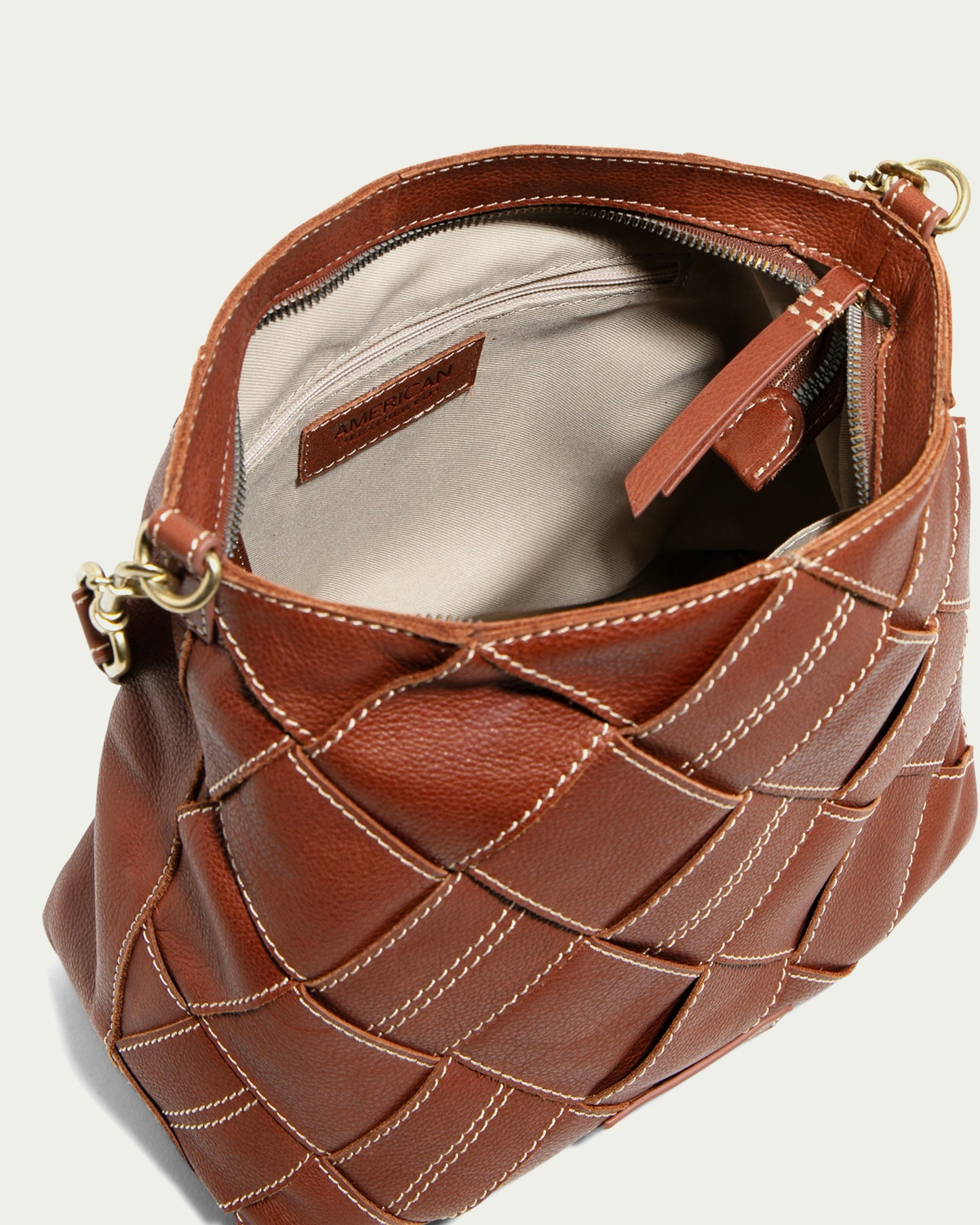 The Mercer Woven Top Zip Hobo by American Leather Co. features brown leather with gold hardware, revealing an open design and a beige interior with a zippered pocket. Its intricate stitching adds elegance, while the removable strap offers versatility to its spacious design.