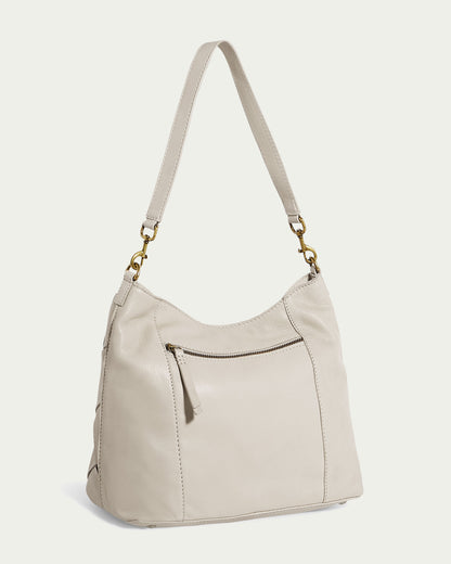 The American Leather Co. Mercer Woven Top Zip Hobo features a cream-colored leather design with a zip-top closure, zippered front pocket, and single strap. Gold hardware connects the strap to the bag, offering an understated look against its plain off-white background.