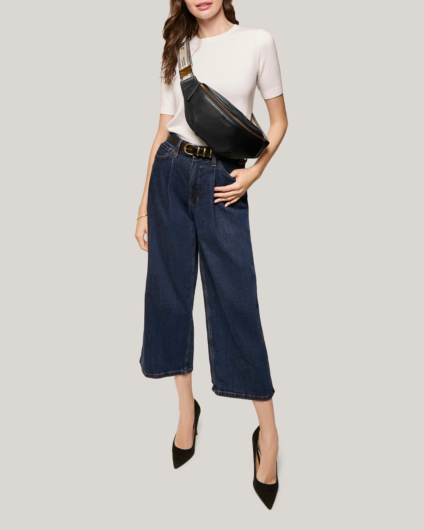 A woman is wearing a white short-sleeve top, dark blue wide-leg cropped jeans, and black high heels. She has an American Leather Co. Millwood Belt Bag with an adjustable webbed strap slung across her chest, perfect for hands-free living. The background is plain light grey.