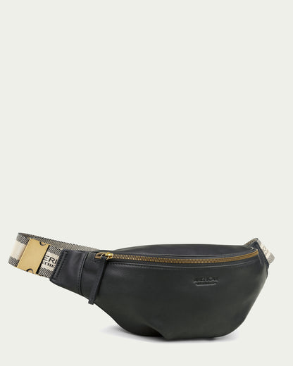 The Millwood Belt Bag by American Leather Co. is a sleek black leather fanny pack accented with a gold zipper. It features an adjustable webbed strap adorned with a contrasting white and black checkered pattern and the word "FRAGILE" in bold black letters on a white background, making it ideal for hands-free convenience. A prominent gold buckle on one side of the strap secures the fit.