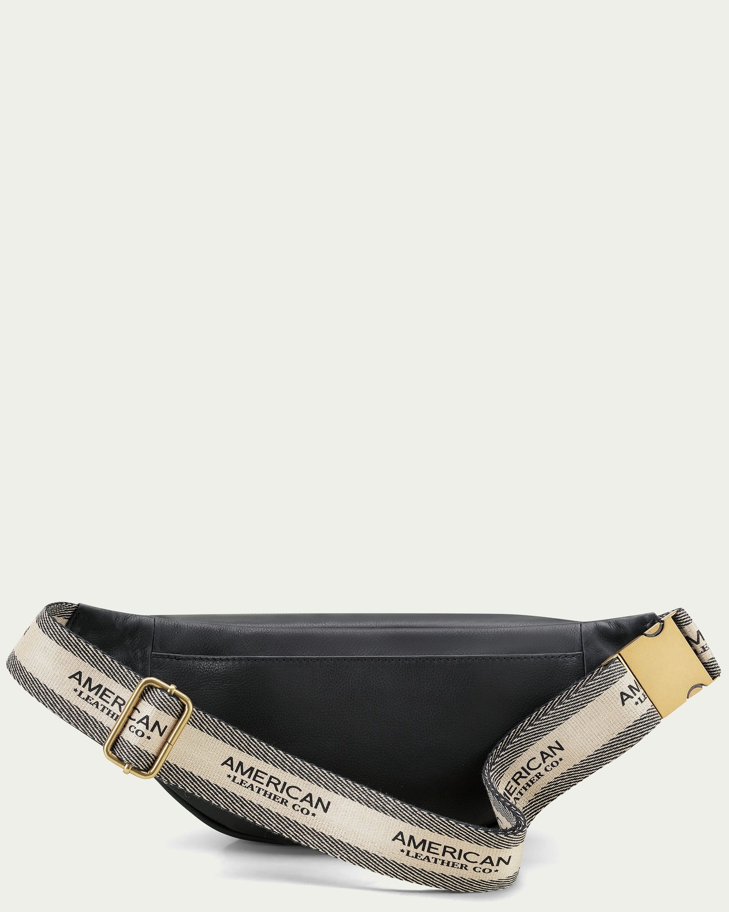 An American Leather Co. Millwood Belt Bag made from black leather, featuring a beige adjustable webbed strap with "AMERICAN LEATHER CO." text in black and a gold metal buckle. Perfect for hands-free living, the bag is set against a plain off-white background.