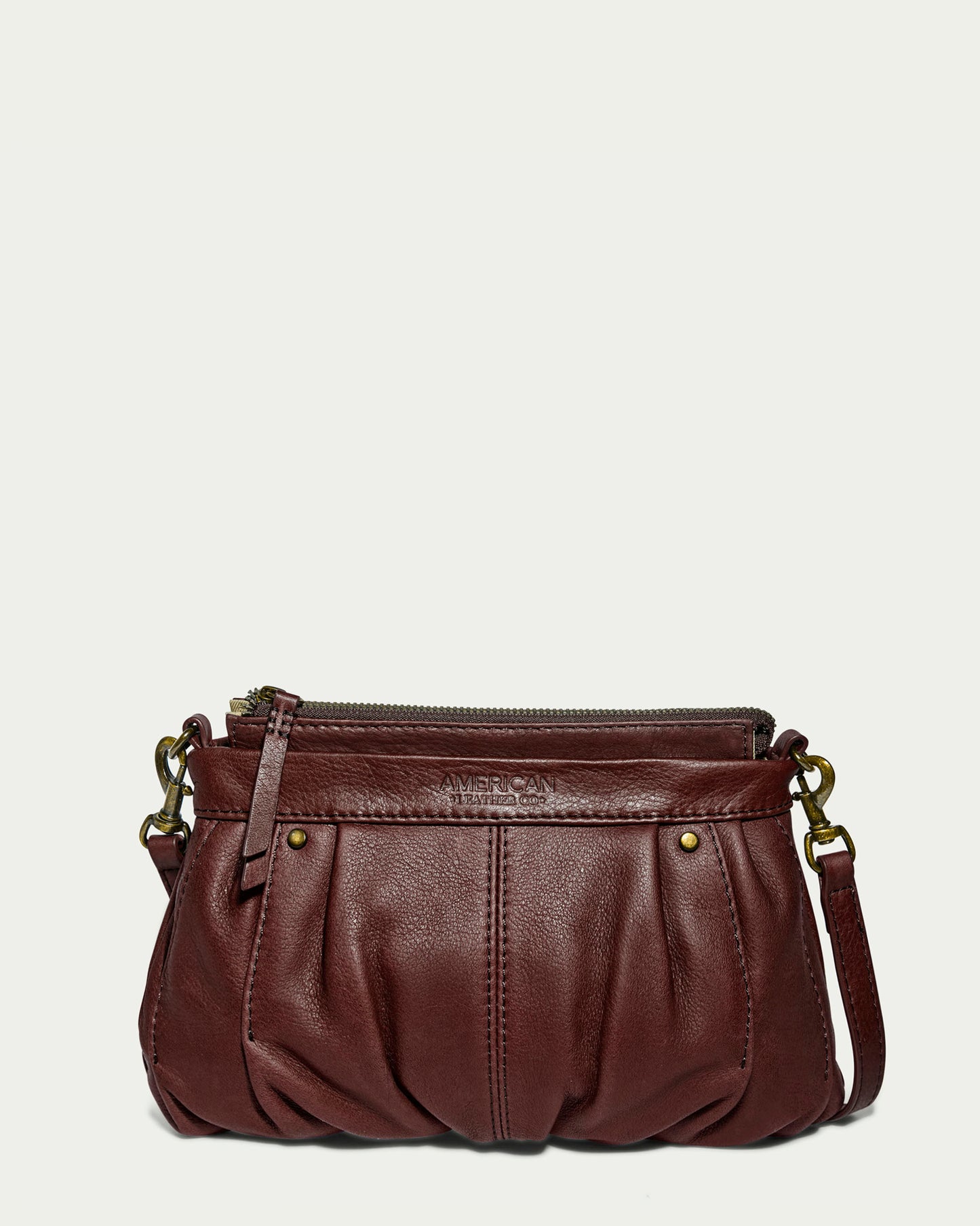 The Saratoga Crossbody by American Leather Co. is a small, stylish bag made from genuine brown glove leather. It features a pleated design on the front, gold-tone hardware, a single zip closure, and an adjustable crossbody strap. The compact style is finished with a subtle embossed brand logo near the top zipper.