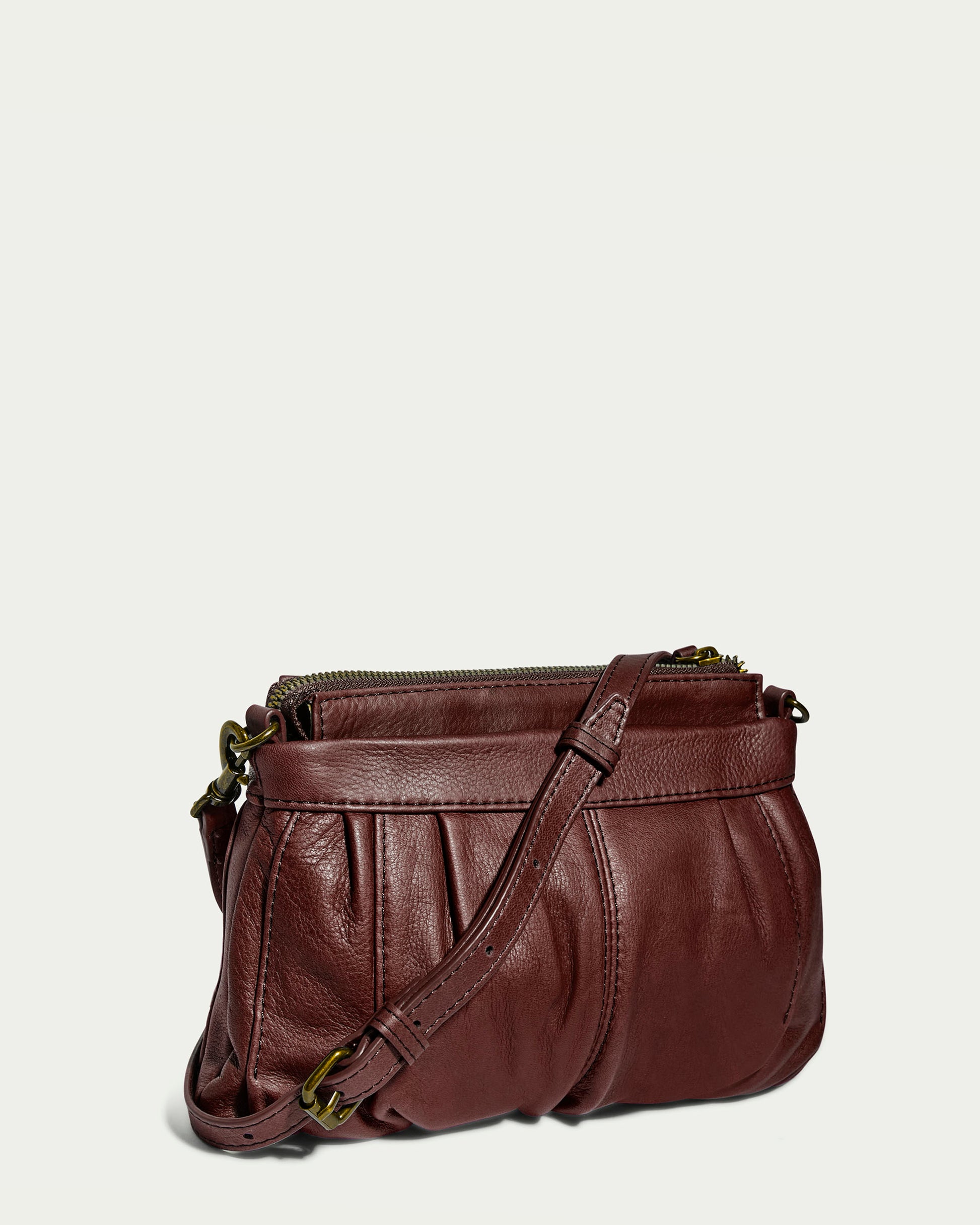 The Saratoga Crossbody by American Leather Co. is a small, stylish burgundy leather purse with a pleated design crafted from genuine glove leather and an adjustable crossbody strap. This purse features gold-tone hardware and a top zip closure, set against a plain, light background.