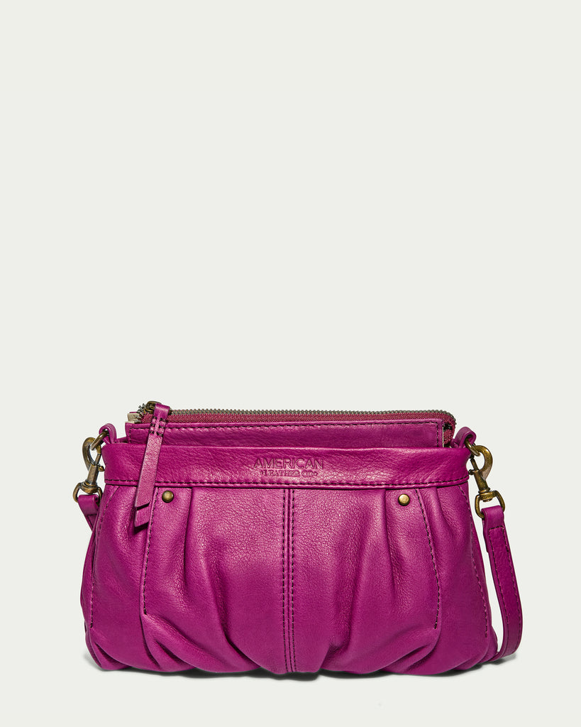 Introducing the Saratoga Crossbody by American Leather Co., a small, vibrant magenta crossbody bag made from genuine leather. It features a soft, ruched design with an adjustable long strap, a secure zipper closure, and decorative brass studs.