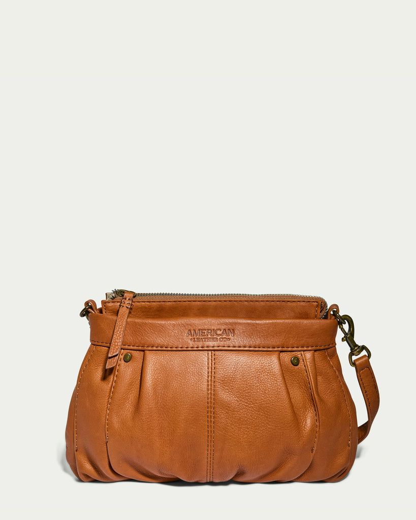 The Saratoga Crossbody from American Leather Co. is a compact leather crossbody purse crafted from genuine glove leather. It features pleated details on the front, two small metal studs, and the brand name "American Leather Co." embossed above. With a zipper closure and an adjustable shoulder strap, this lightweight essential looks perfect against a light, neutral background.