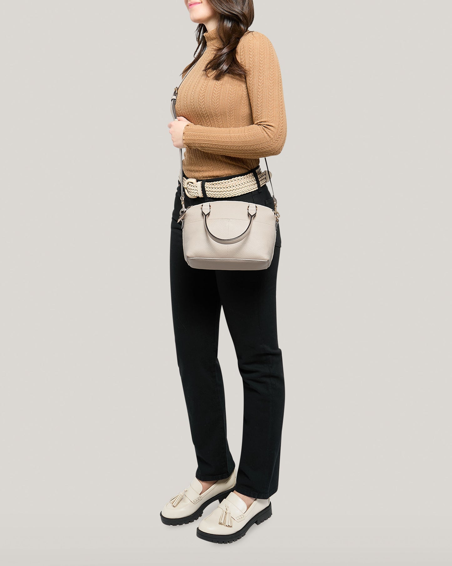 A woman stands against a plain background wearing a brown sweater and black pants, carrying the Carrie Mini Dome Crossbody from American Leather Co., featuring an adjustable strap. With beige tassel loafers matching her style, her long hair elegantly rests on her shoulders.