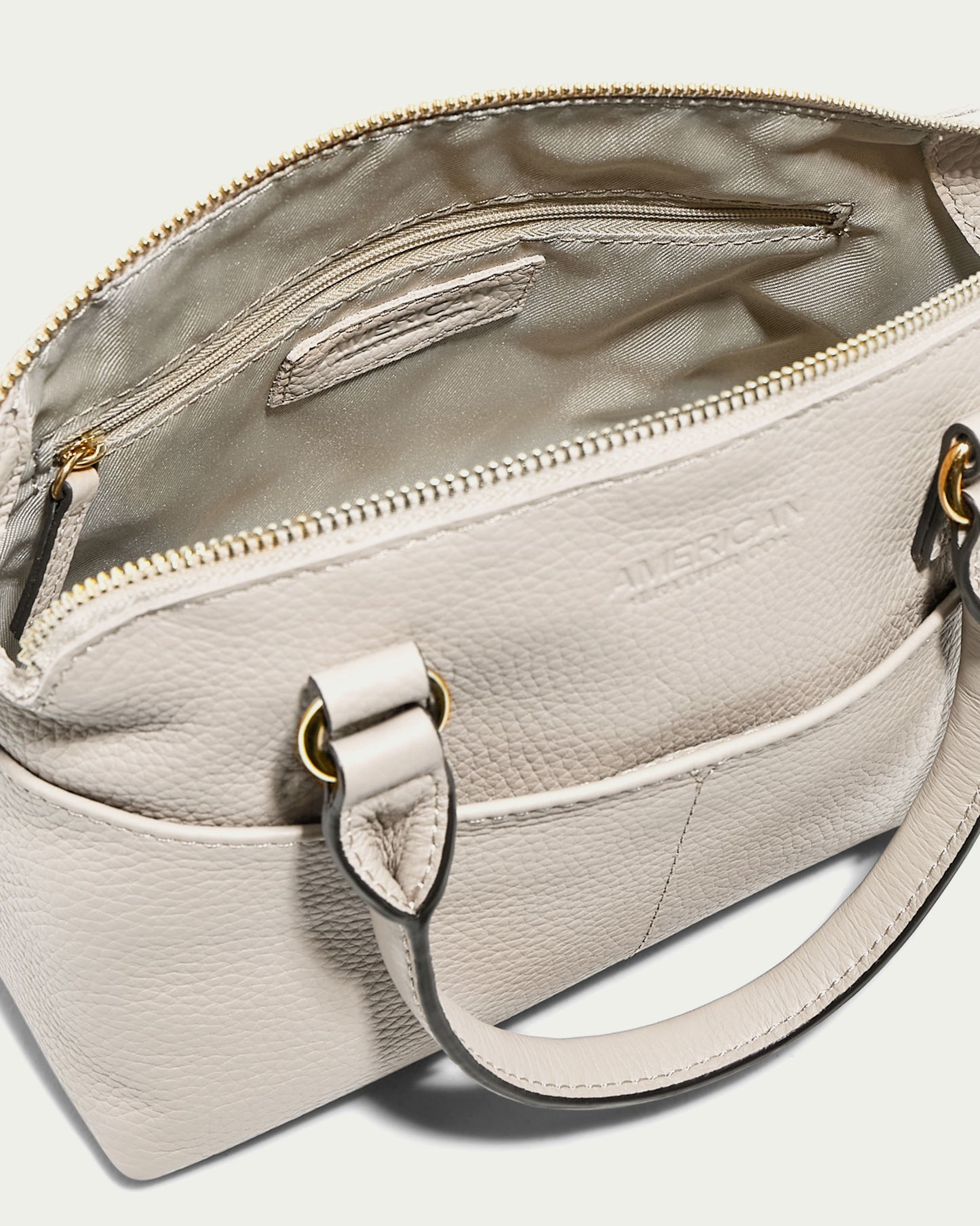 The Carrie Mini Dome Crossbody by American Leather Co. is crafted from genuine white leather and has gold hardware with a zippered opening. It features a light fabric lining, an interior zip pocket, an embossed brand name on the exterior, and an adjustable crossbody strap for versatile wear.