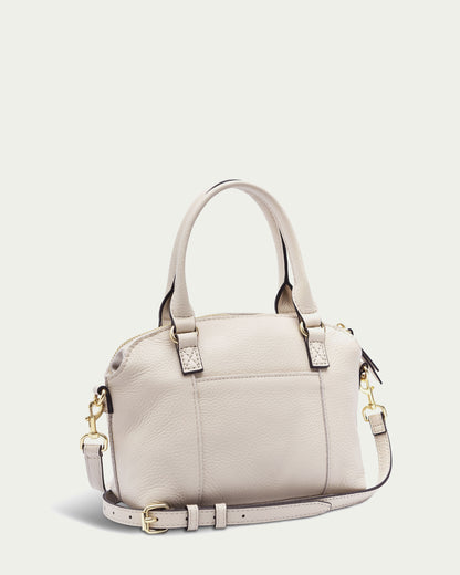 The Carrie Mini Dome Crossbody by American Leather Co. is a beige leather handbag with two short handles and a detachable, adjustable crossbody strap. It features subtle gold hardware and a minimalist design against a plain, light background.