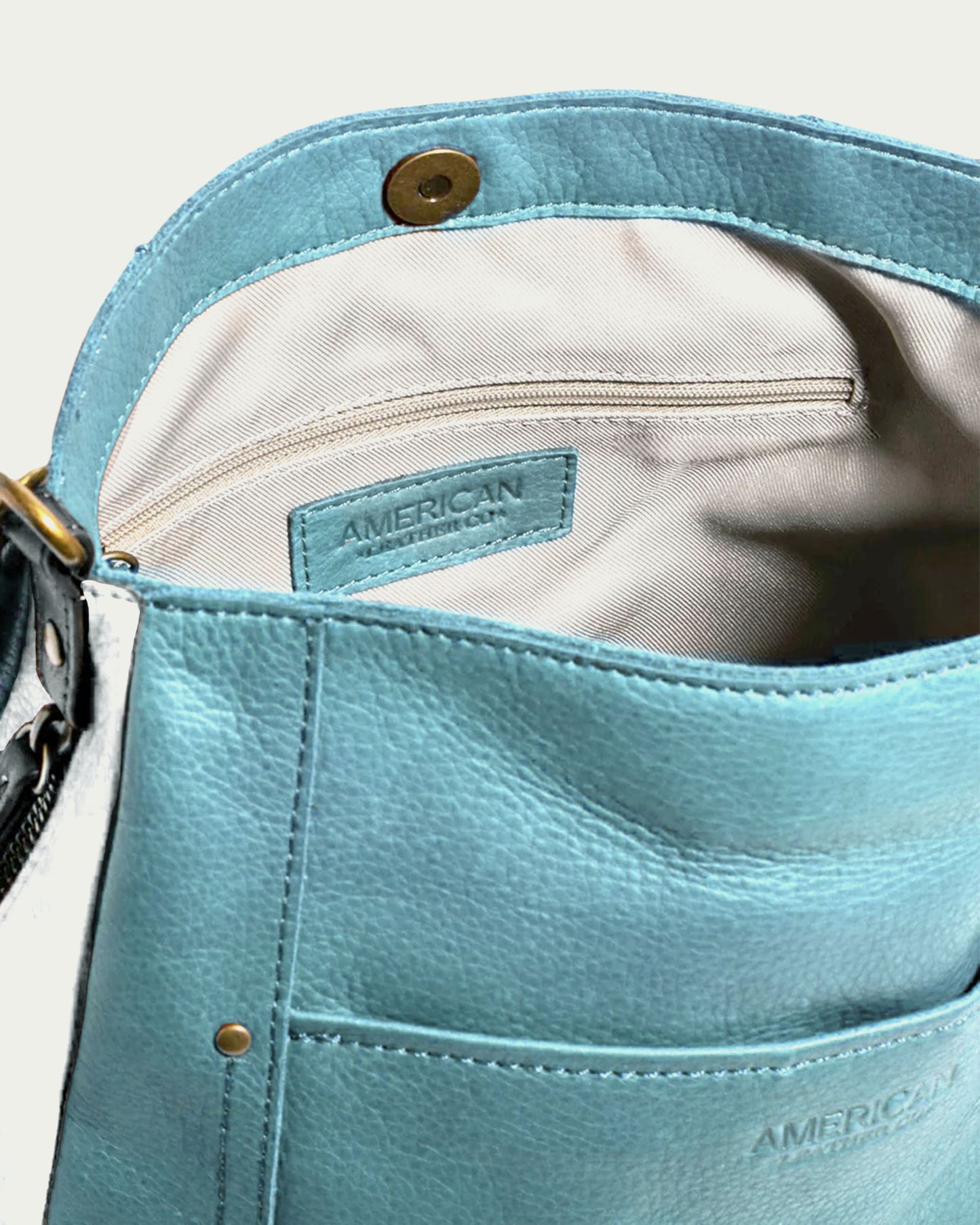 This Austin Shoulder bag from American Leather Co. is crafted from genuine American leather and boasts a light blue tone. It features a visible interior zipper pocket and a gold snap button closure, with an "American" label sewn onto the lining, making it an ideal casual accessory.