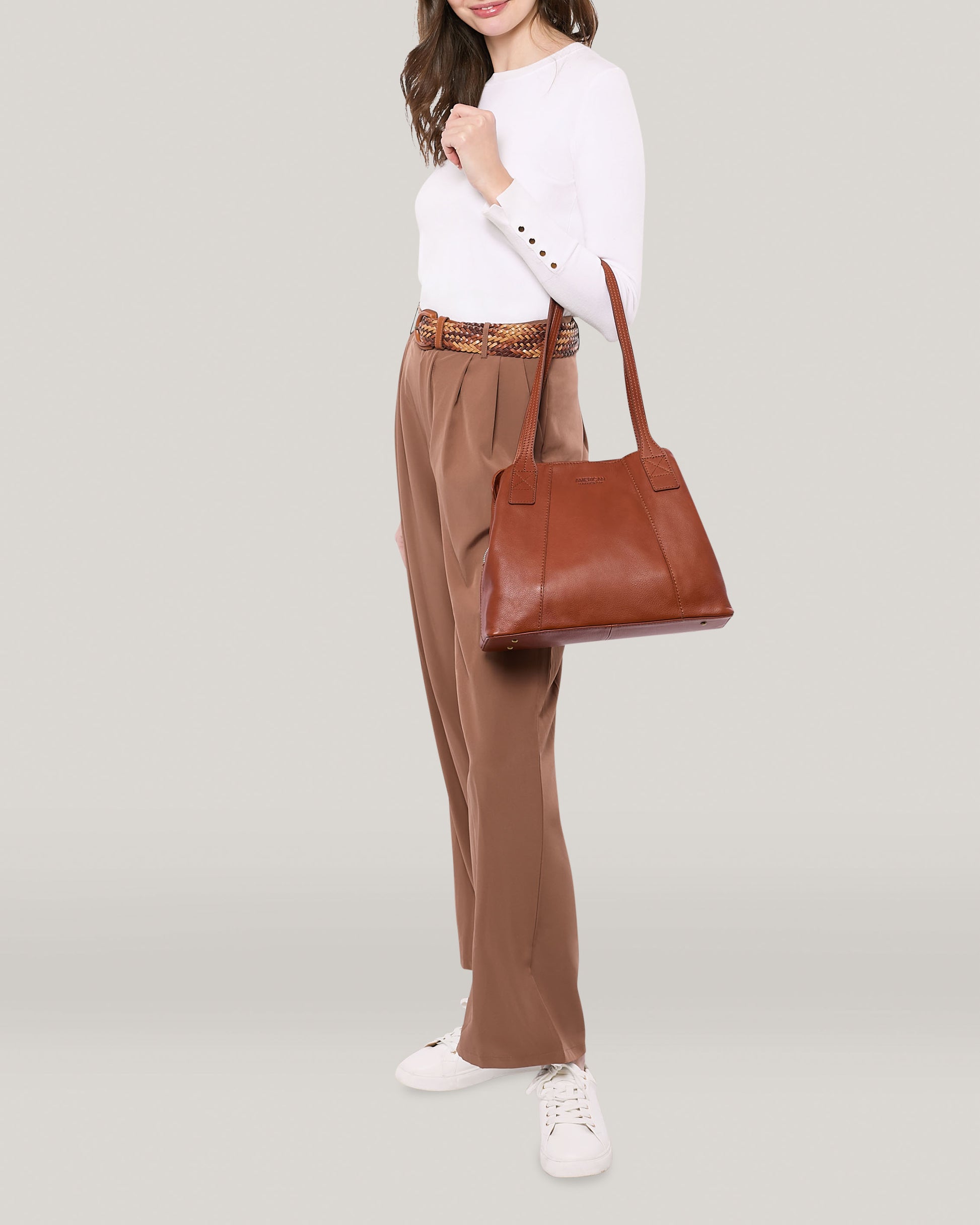 A person is standing against a plain background, dressed in a white long-sleeve top and brown trousers, accessorized with a brown belt. They are carrying the Ada Triple Entry Tote by American Leather Co., made from genuine leather, and wearing white sneakers. Their face is partially visible to the left.