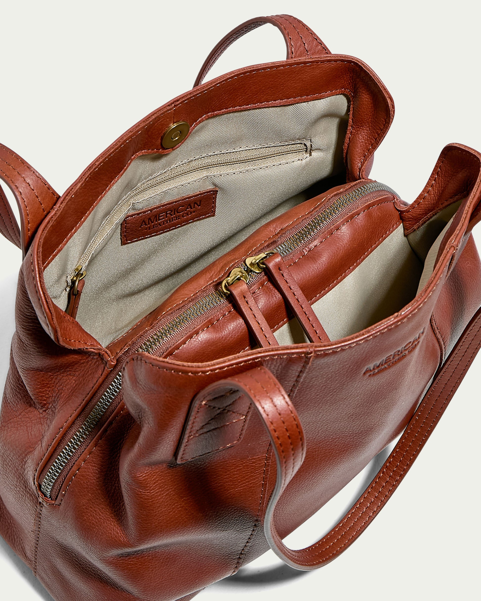 A top view of the open Ada Triple Entry Tote by American Leather Co. showcases its rust-brown genuine leather exterior and well-organized interior. The beige lining features multiple compartments, including a zippered pocket and a visible brand label, highlighting excellent interior organization. The tote is equipped with both zipper and snap closures, and includes two sturdy handles.