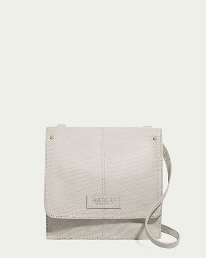 The Brook Flap Crossbody by American Leather Co. is a small, light gray textured leather bag with subtle stitching and an embossed brand name on the front. It features genuine leather, an adjustable strap for versatile wear, combining style and function.