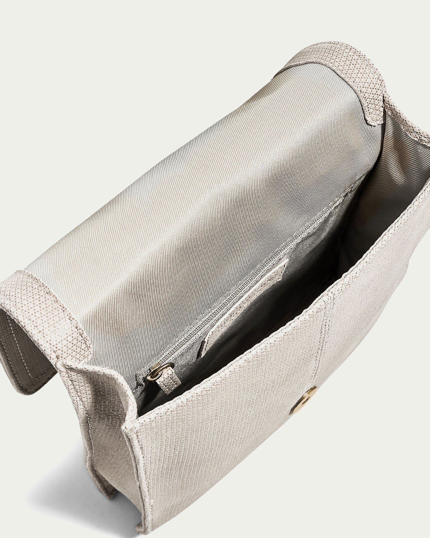 The American Leather Co. Brook Flap Crossbody, an open beige fabric purse, unveils its inner lining and zippered pocket. It features a simple, elegant design with a button clasp and an adjustable strap for versatile wear.