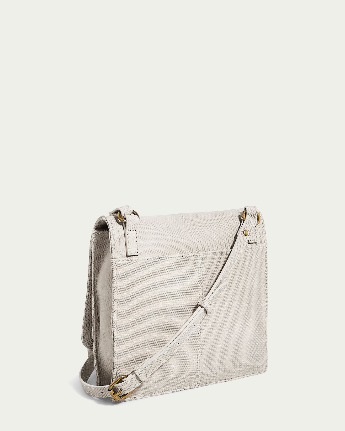 The American Leather Co. Brook Flap Crossbody is a beige, genuine leather bag with a textured look on a light background. It has an adjustable strap with gold-tone buckles and a structured, rectangular flap closure.