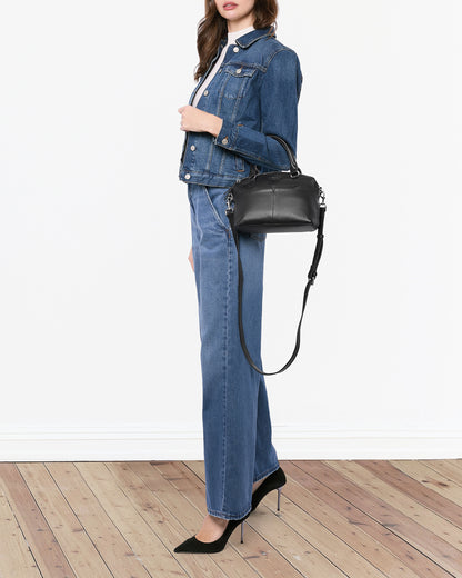 A woman stands against a white background on a wooden floor, wearing a denim jacket over a white top and high-waisted, wide-leg denim jeans. She holds an American Leather Co. Carrie Mini Dome Crossbody with an adjustable strap in one hand and wears black pointed-toe heels.