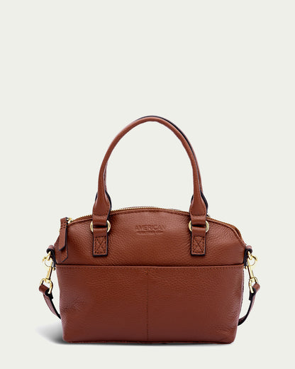 A Brandy Pebble leather handbag with two short handles and gold-tone hardware, this Genuine Leather Carrie Mini Dome Crossbody from American Leather Co. features an exterior pocket on the front, a top zipper closure, and "AMERICAN LEATHER CO." embossed on the front. An adjustable strap adds versatility. Placed against a light background.