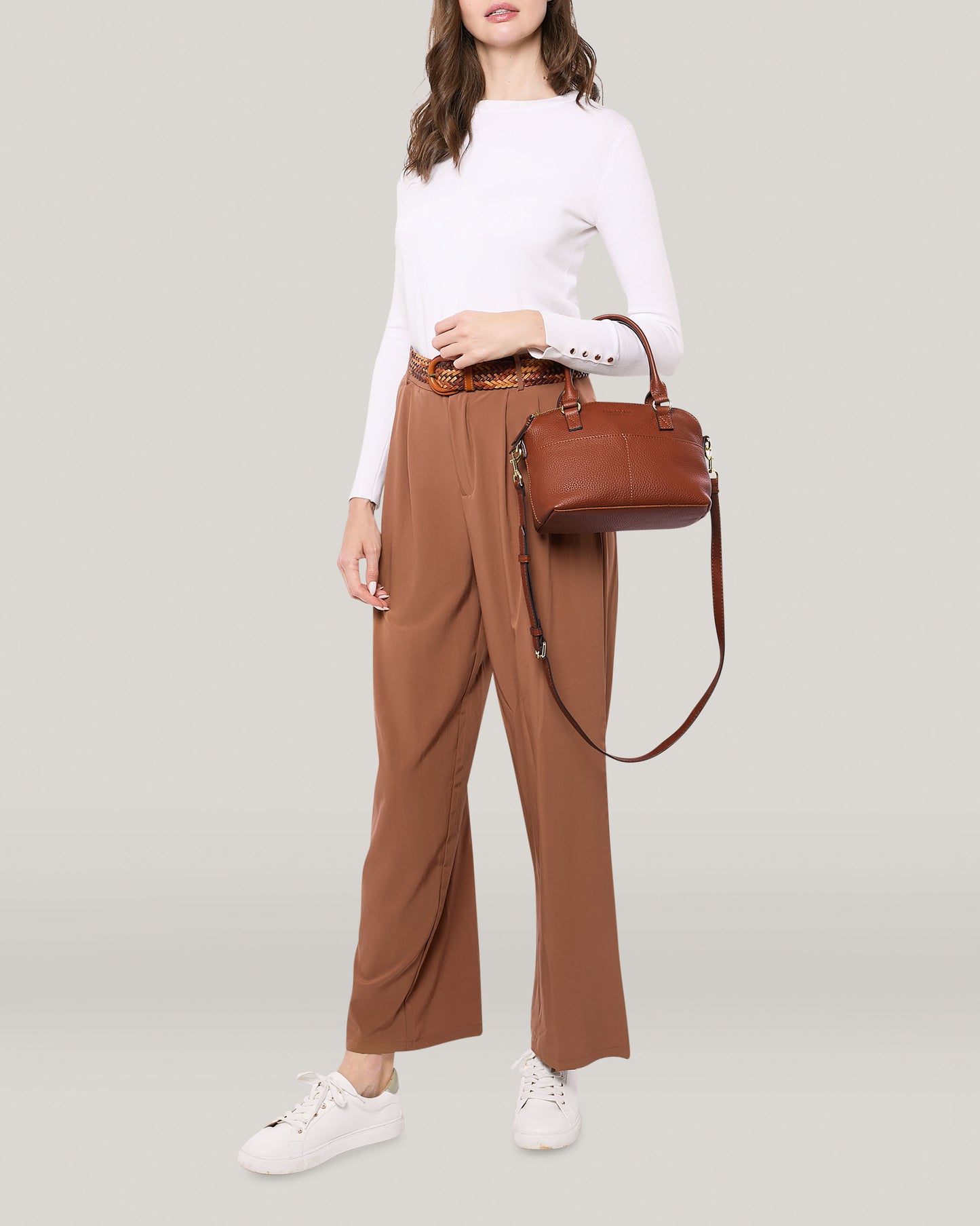 A person wearing a white long-sleeve shirt and high-waisted, loose brown pants with a brown belt is holding a Carrie Mini Dome Crossbody by American Leather Co., crafted from genuine leather. They are wearing white sneakers against a neutral, light gray background, with their face partially obscured.