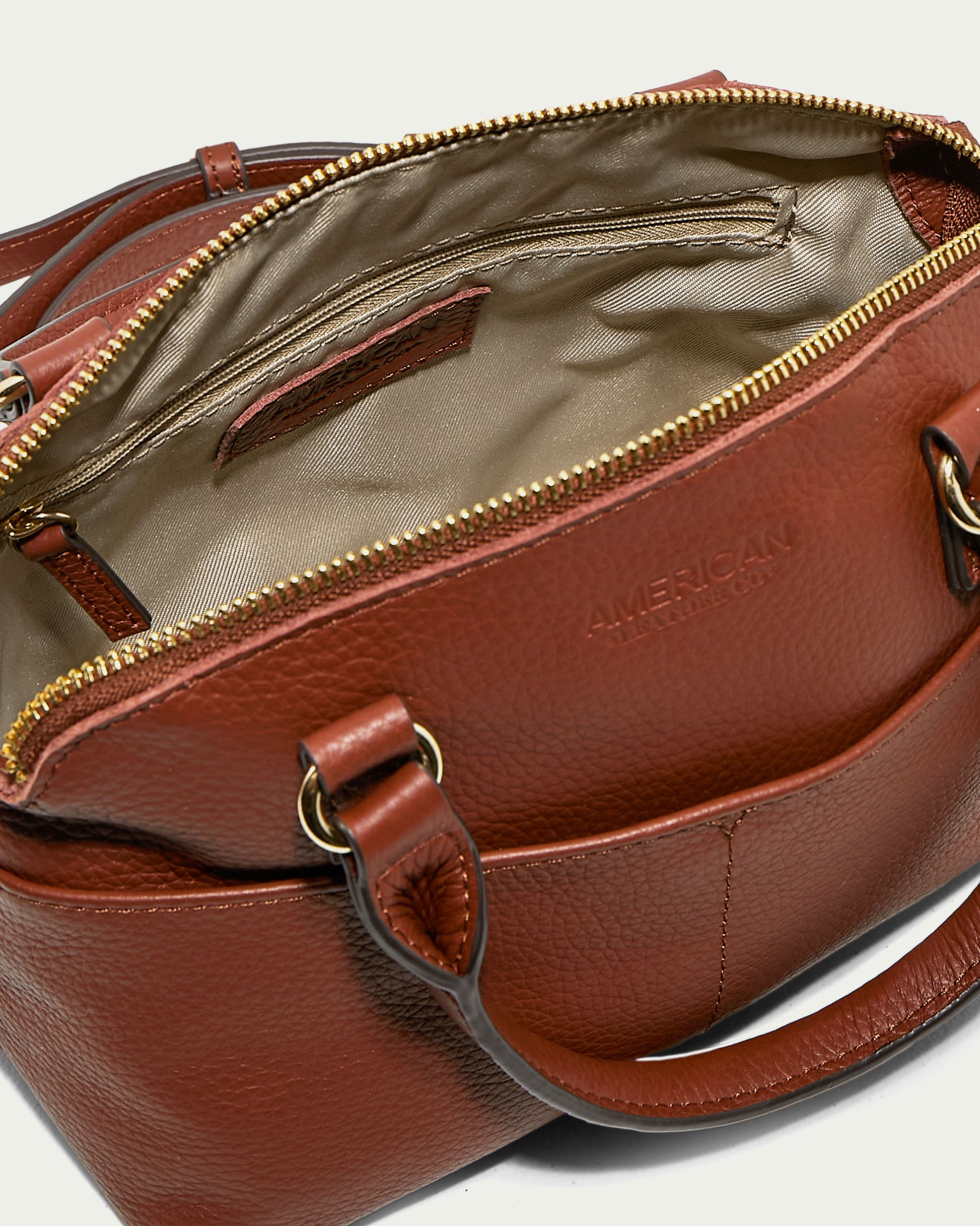 The Carrie Mini Dome Crossbody from American Leather Co. is open, showcasing a beige interior lining and an inside zipper pocket. The brand name "American Leather Co." is embossed on the exterior of this brown leather handbag, which boasts a top zipper closure. This genuine leather bag features dual handles, gold-toned hardware, and an adjustable strap.