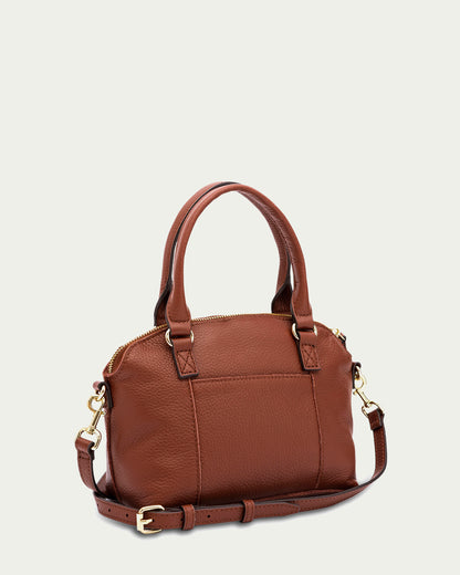 A stylish brown leather handbag with gold hardware, the Carrie Mini Dome Crossbody by American Leather Co. features dual top handles and an adjustable shoulder strap with a buckle. The bag boasts a zippered main compartment and an exterior pocket, displayed against a plain light-colored background.