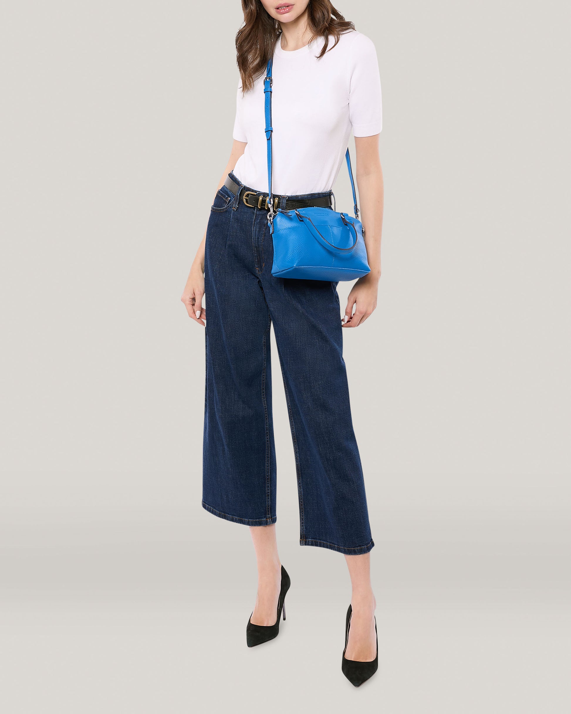 Person standing against a plain background wearing a white short-sleeve top, dark wide-leg jeans, and black heels. They have the American Leather Co. Carrie Mini Dome Crossbody, a blue genuine leather bag with multiple pockets, over their shoulder. The person's face is not visible.