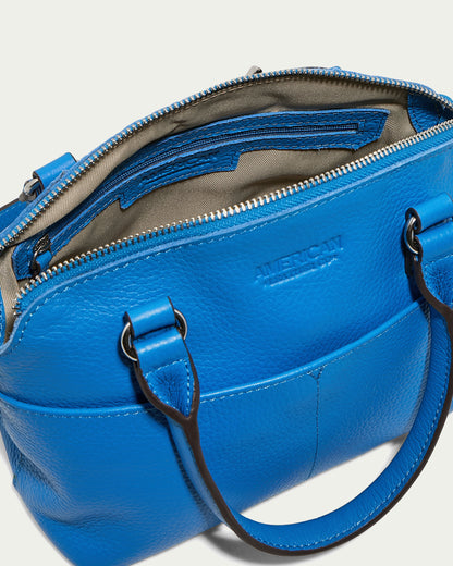 An open blue Carrie Mini Dome Crossbody by American Leather Co. made of genuine leather and equipped with dual straps. It boasts multiple compartments, including a zippered pocket on the inner side, while its beige fabric lining adds a touch of elegance. The outer side is stylishly embossed with the word "AMERICAN.