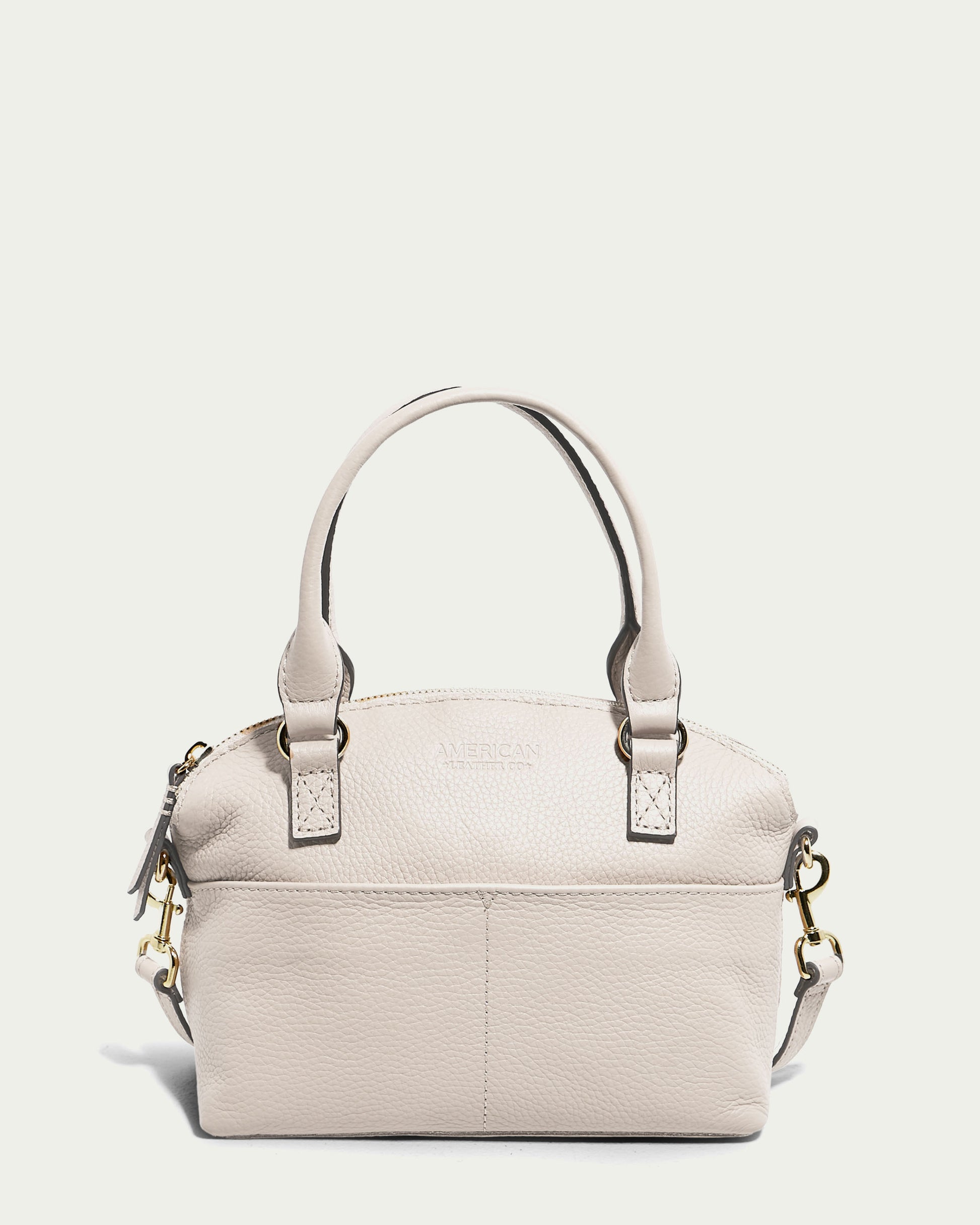 An ecru pebble leather handbag with two handles and an adjustable shoulder strap is displayed against a plain white background. The Carrie Mini Dome Crossbody from American Leather Co. features a textured finish, gold hardware, and a front pocket, with the brand name subtly embossed on the front.