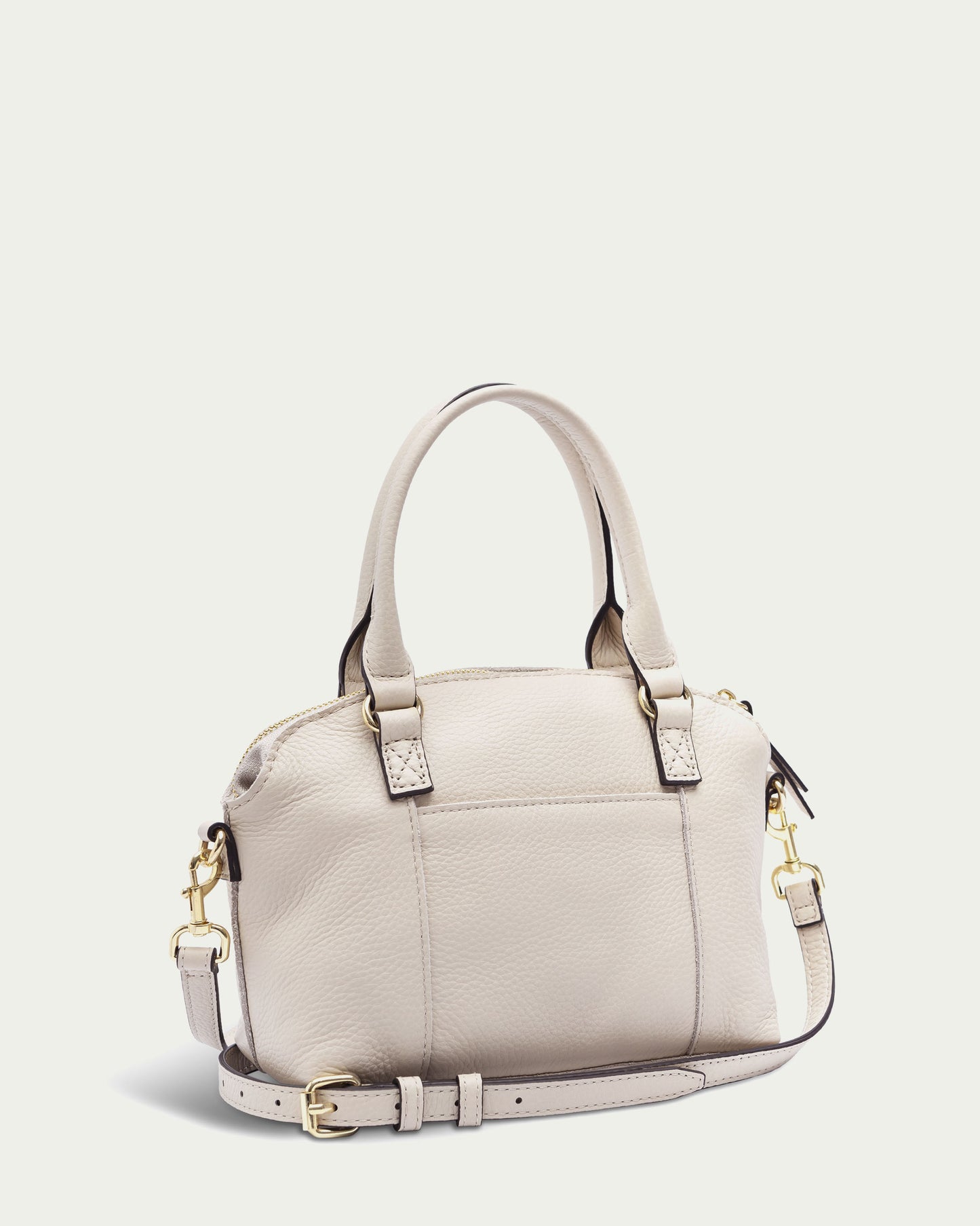 A chic beige handbag from American Leather Co., the Carrie Mini Dome Crossbody, is showcased. It boasts two hand-carry straps and a detachable, adjustable shoulder strap. The bag includes a zippered main compartment, gold hardware, and a minimalist design that exudes elegance.