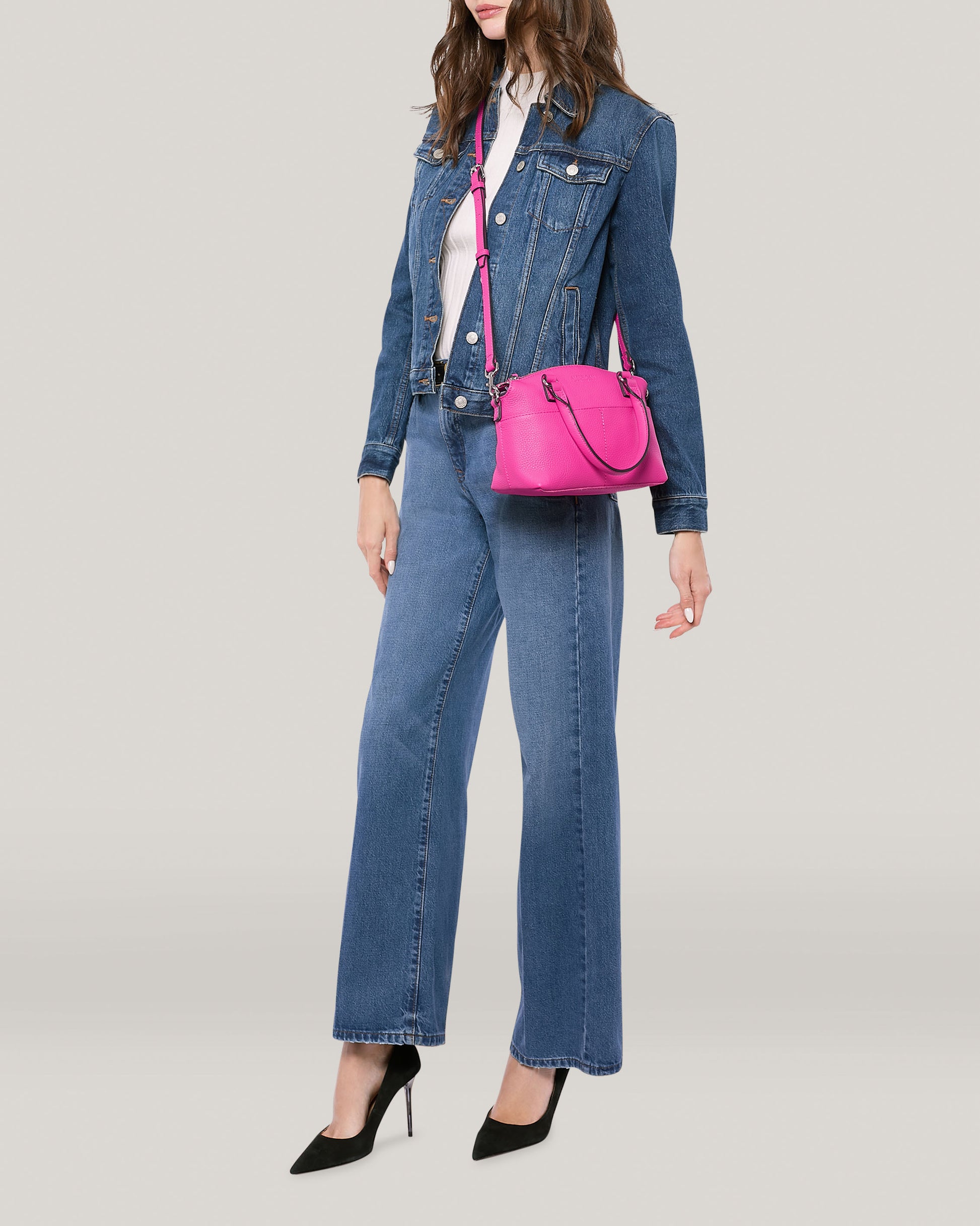 A person is wearing a blue denim jacket paired with matching denim jeans and a white shirt. They have the Carrie Mini Dome Crossbody by American Leather Co., which is bright pink, slung over their shoulder, and are complemented by black high-heeled shoes. The background is plain and light-colored.