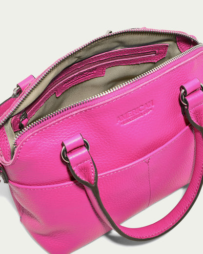 The Carrie Mini Dome Crossbody from American Leather Co. is shown in a vivid pink color with its top zippered compartment partially open, revealing an interior lined with olive green fabric. The women's crossbody bag features two sturdy handles and a front pocket, with the word "American" embossed on the exterior.