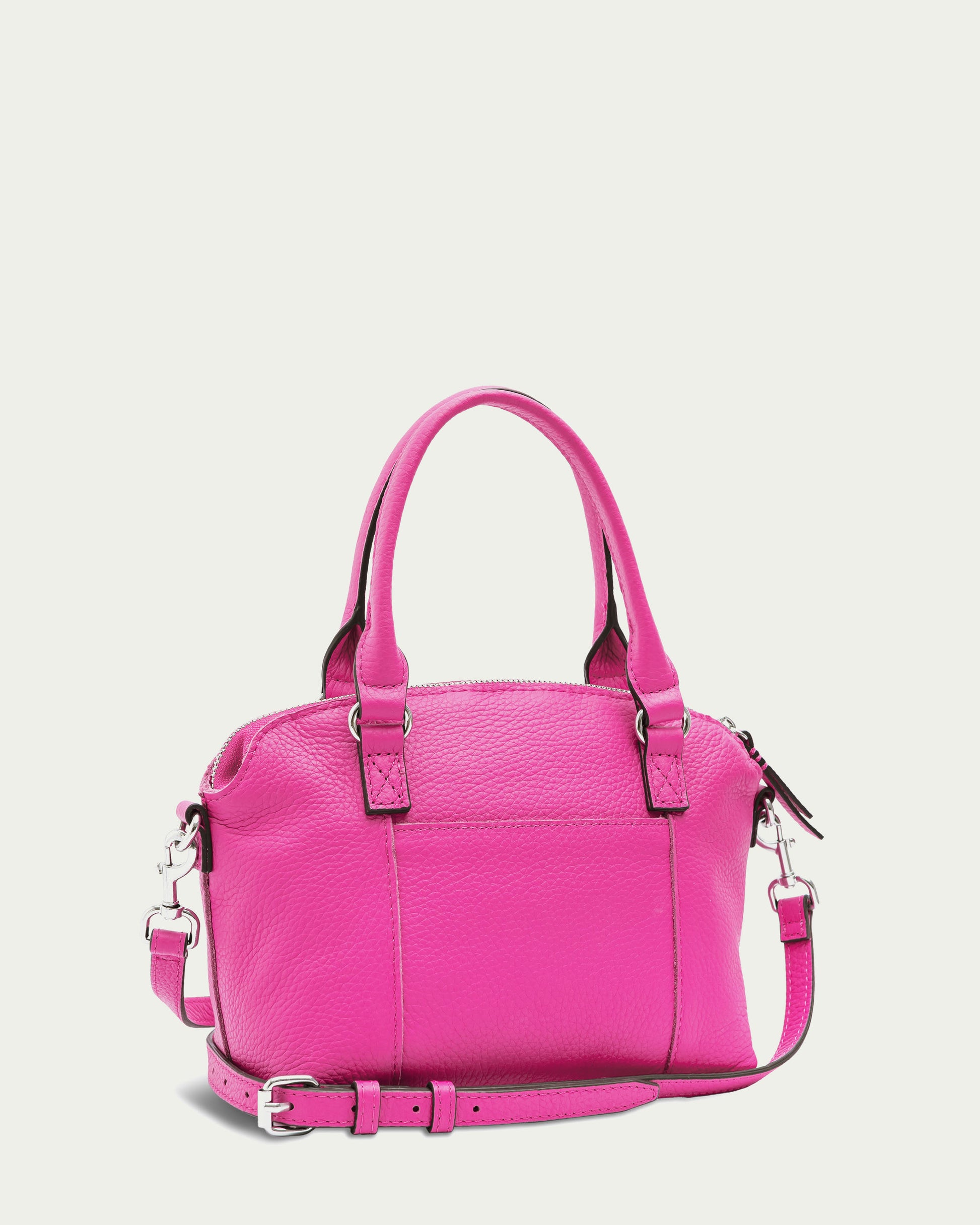 The Carrie Mini Dome Crossbody by American Leather Co. is a bright pink leather bag featuring a pebbled texture. It comes with two handles and a detachable, adjustable shoulder strap adorned with silver hardware accents, including buckles and clasps on the strap and zipper pull. The background is white.