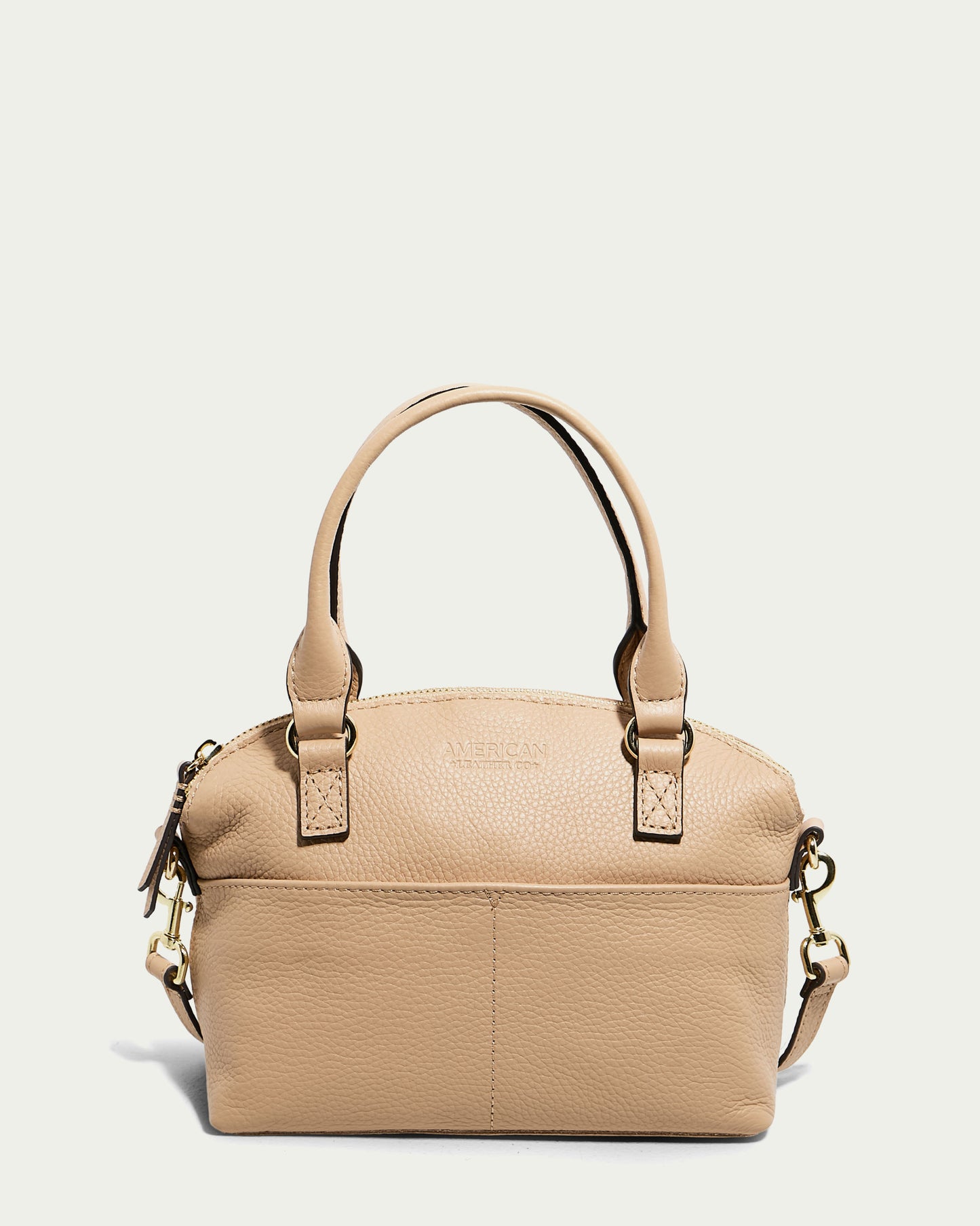 The Carrie Mini Dome Crossbody by American Leather Co. is a macadamia pebble-textured leather women's crossbody bag. It features two short handles, gold-toned hardware, a top zipper closure, and a small front pocket. "American" is embossed on the front of the bag, which also includes a detachable shoulder strap on the side.