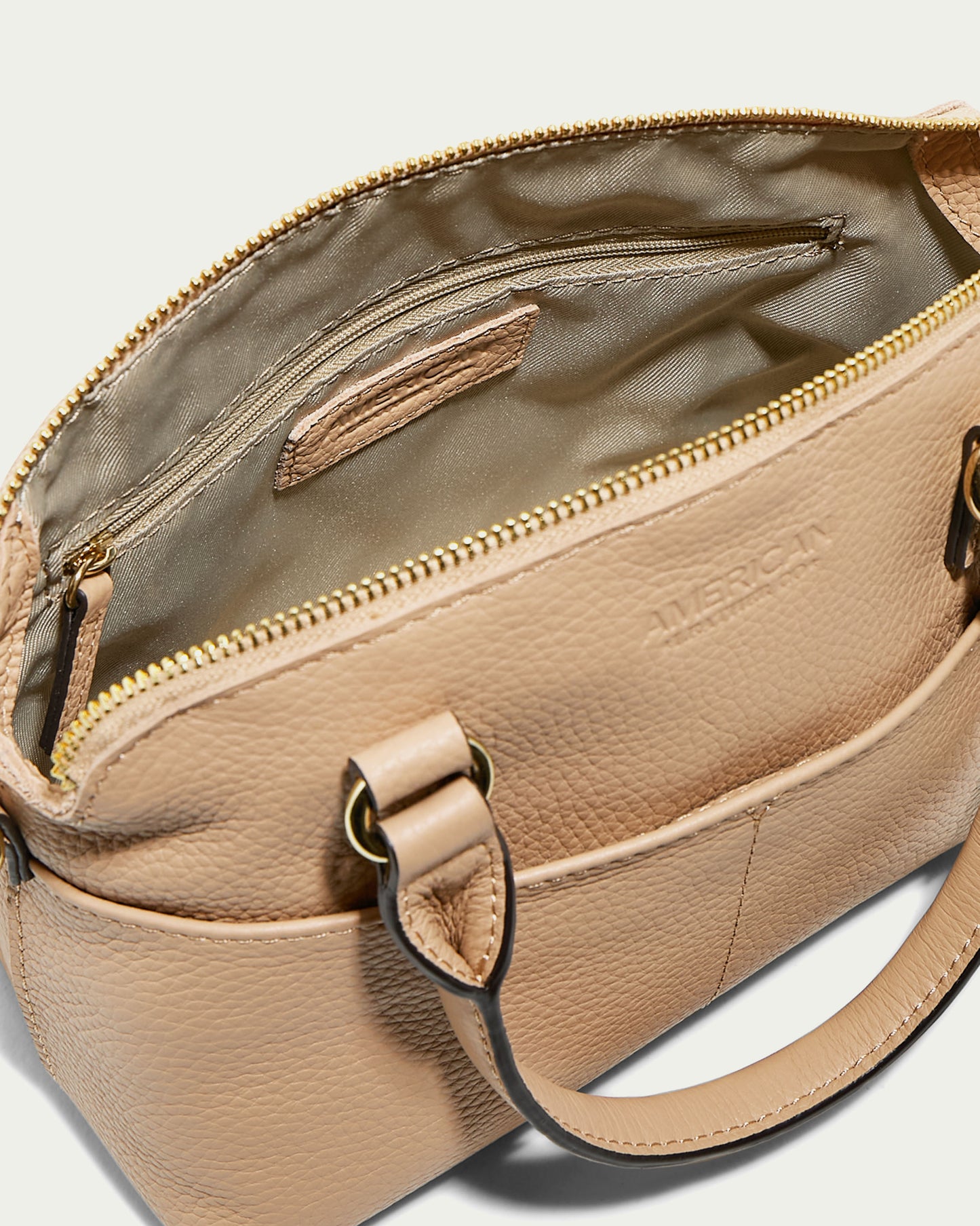 The Carrie Mini Dome Crossbody from American Leather Co., crafted from beige genuine leather, features a top zipper and opens to reveal a lined interior. This elegant women's crossbody bag includes a handle and metal clasps for an optional shoulder strap. The word "AMERICAN" is embossed on the front side of the bag.