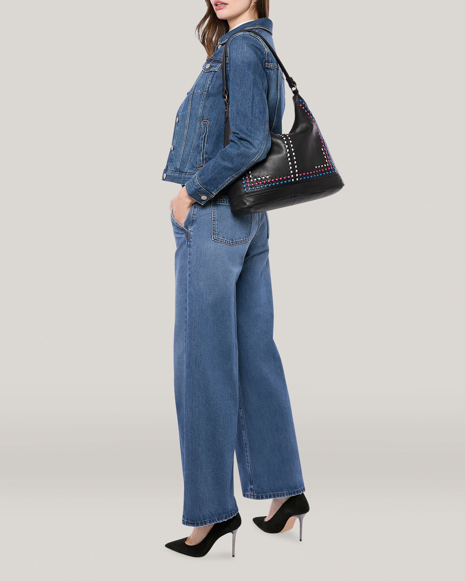 A woman, dressed in a denim jacket and wide-leg jeans, is standing in profile against a plain light gray background. She has an Evelyn Hobo bag from American Leather Co., featuring a studded design, slung over her shoulder and is wearing black high-heeled shoes.