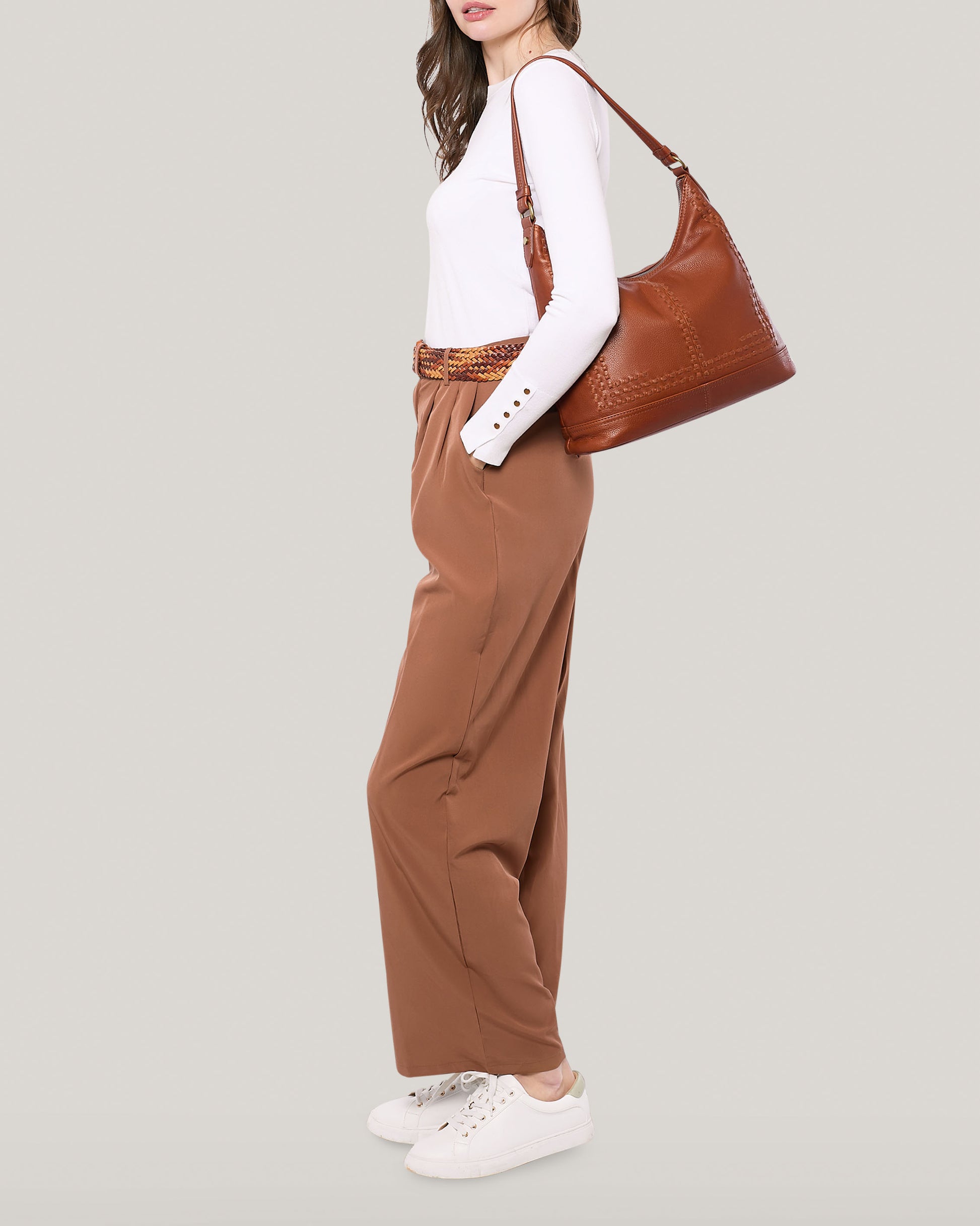 A woman is standing against a light grey background wearing a white long-sleeve top, high-waisted brown pants, and white sneakers. She is holding an Evelyn Hobo shoulder bag from American Leather Co. Her brown hair is styled loosely and falls over her shoulder.